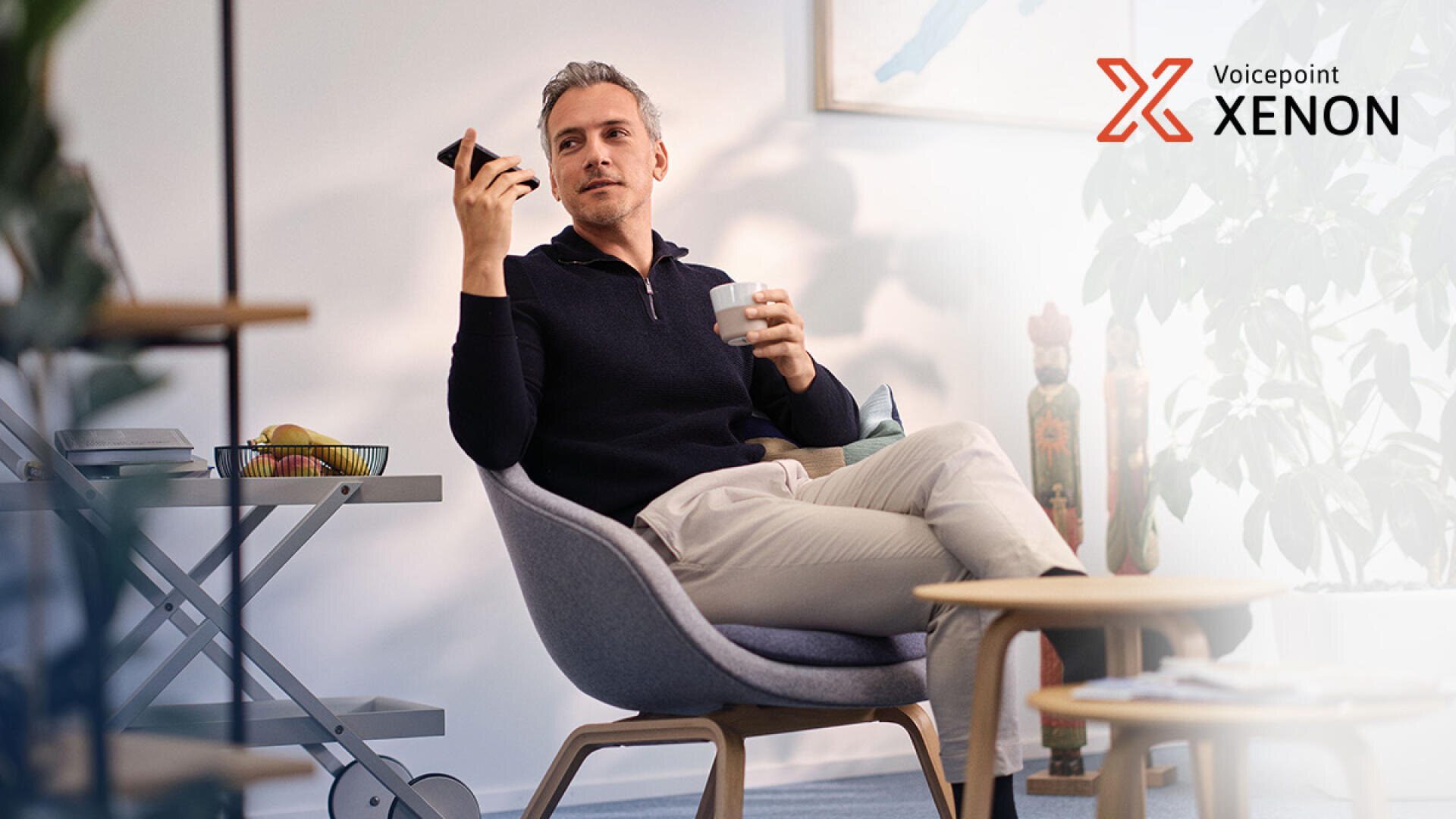 Ein Mann sitzt entspannt auf einem modernen Stuhl und hält ein Smartphone sowie eine Tasse in der Hand. Der Raum ist minimalistisch eingerichtet und mit Zimmerpflanzen sowie einer Obstschale ausgestattet. In der oberen rechten Ecke ist das Voicepoint-Xenon-Logo zu sehen.