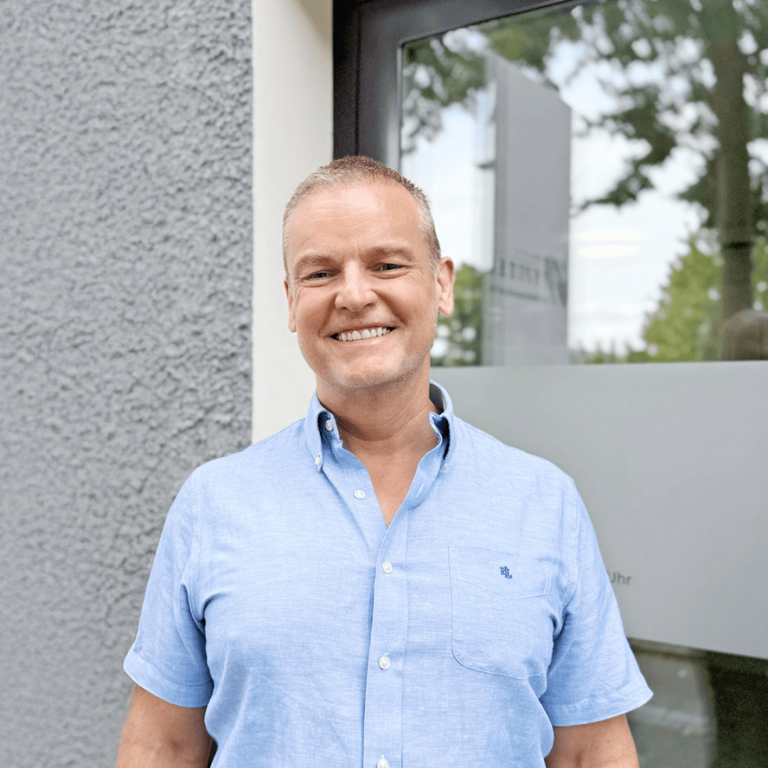 Ein lächelnder Mann in einem hellblauen Kurzarmhemd steht draußen vor einer strukturierten grauen Wand und einer Glastür, hinter ihm spiegeln sich Bäume und der Himmel im Fenster.
