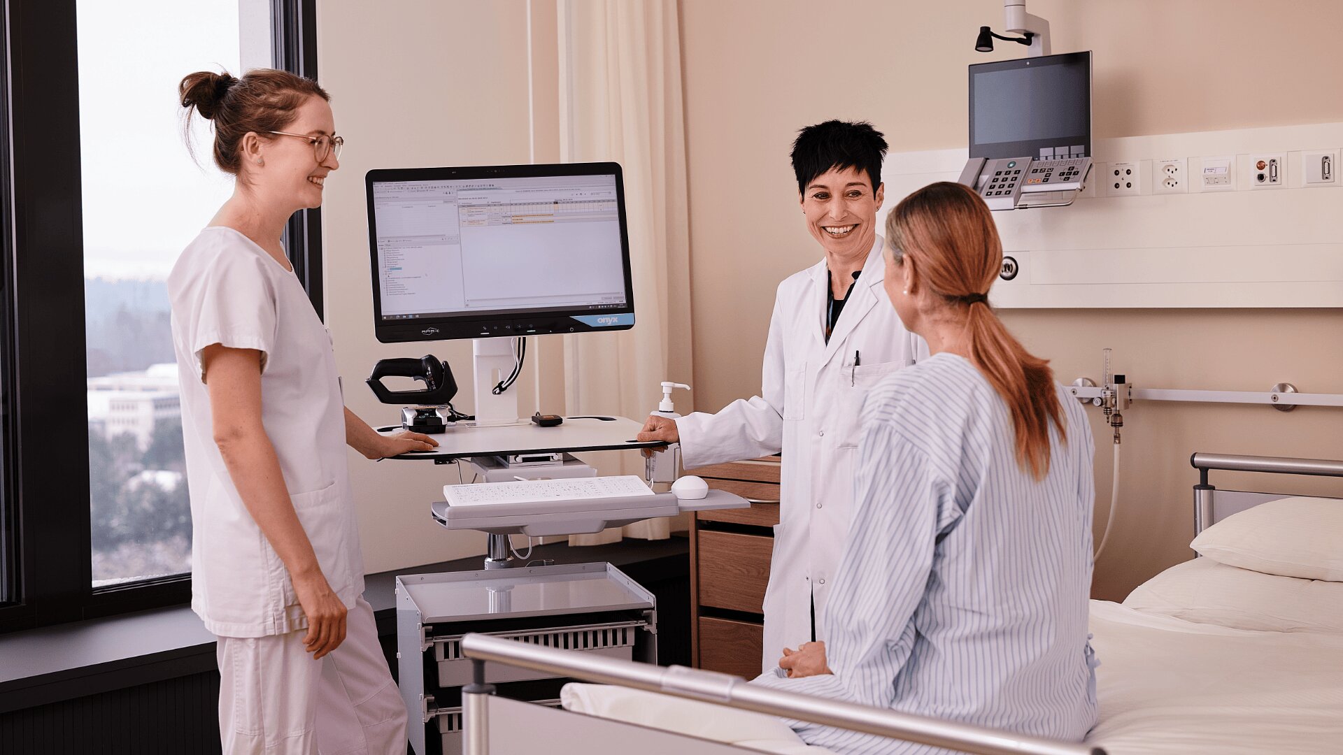 Ein Arzt und eine Krankenschwester stehen an einem Krankenhausbett, lächeln und sprechen mit einem Patienten, der aufrecht sitzt. Ein Computer auf einem mobilen Arbeitsplatz zeigt die medizinischen Daten an. Der Raum ist hell und modern.