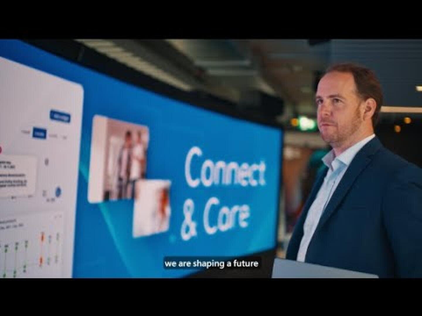 Un homme en costume se tient devant un grand écran numérique sur lequel apparaissent les mots « Connect & Care ». La légende en bas de l'image indique : « Nous façonnons l'avenir. ».