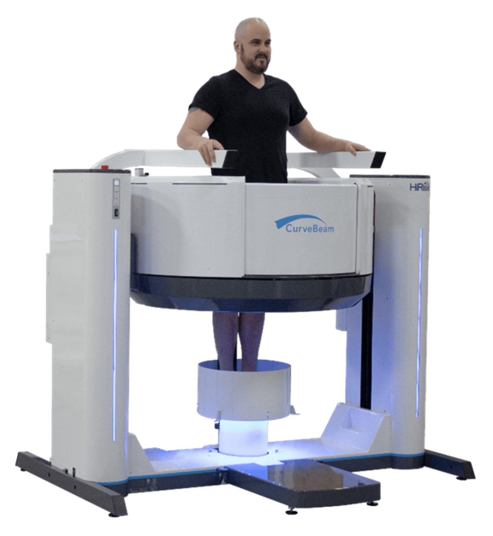 Un homme se tient debout dans un appareil d'imagerie médicale CurveBeam, les pieds à l'intérieur de l'appareil et les mains posées sur des repose-mains. L'appareil émet une lumière bleue et présente un design blanc et moderne.