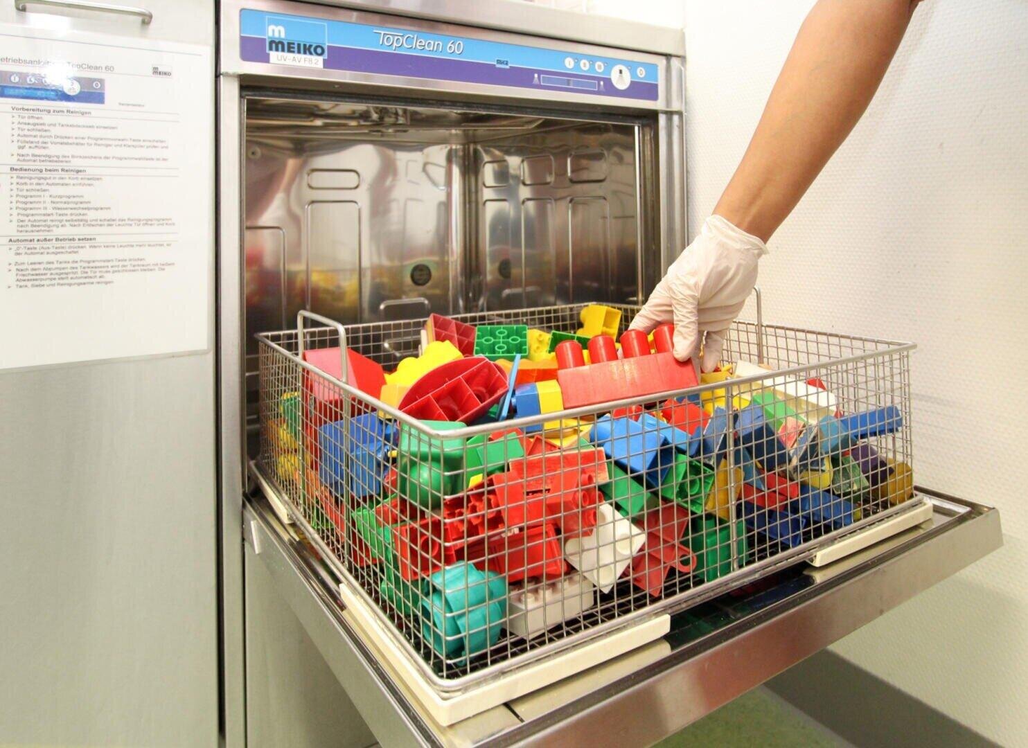 Eine behandschuhte Hand stellt einen Korb voller bunter Plastikspielzeuge in ein spezielles Spülgerät der Marke MEIKO, Modell TopClean 60.