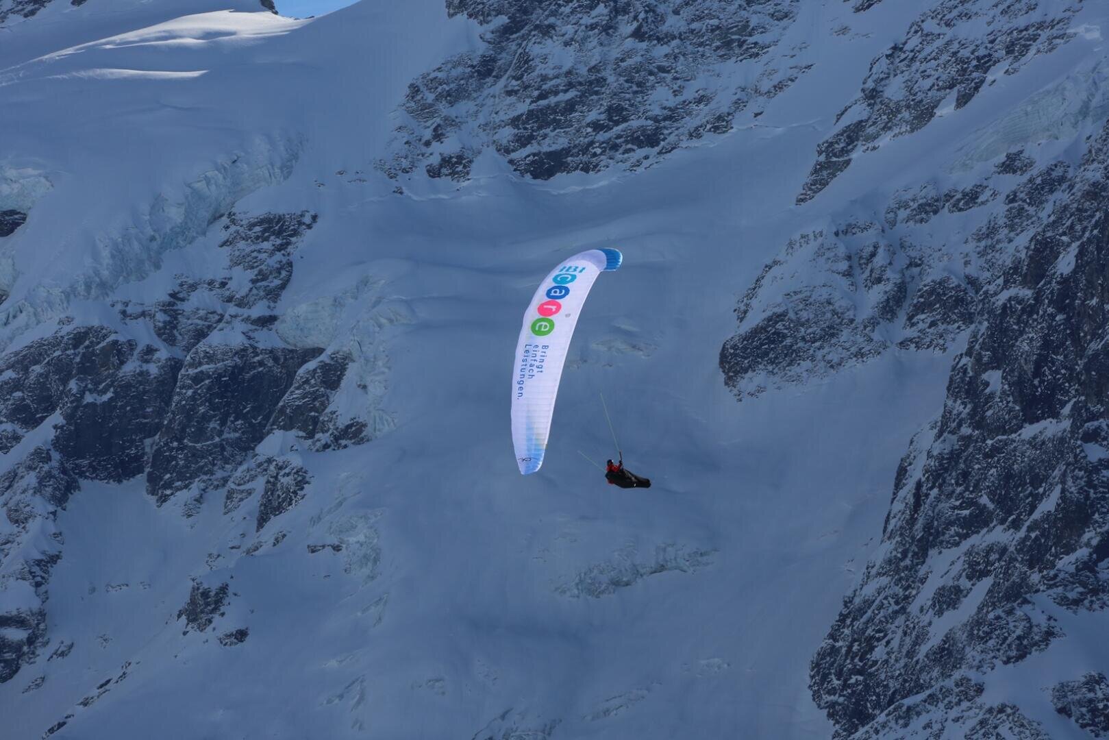 Ein Gleitschirm mit einem bunten Flügel schwebt im strahlenden Sonnenlicht in der Nähe von schneebedeckten, schroffen Bergklippen.