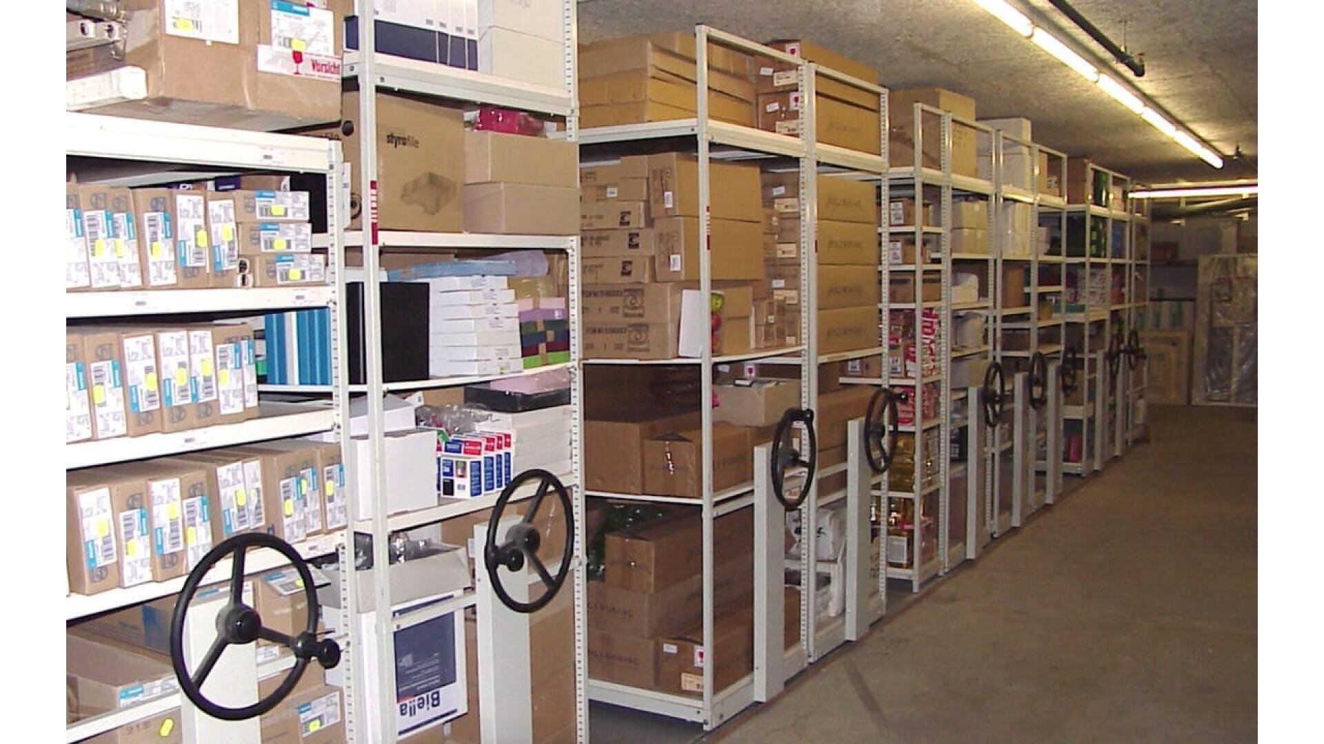 Des étagères remplies de boîtes, de classeurs et de dossiers sont organisées dans une salle de stockage. Certaines étagères sont munies de grandes poignées tournantes noires qui permettent de déplacer les unités. La pièce est éclairée par des plafonniers et le sol est en béton.