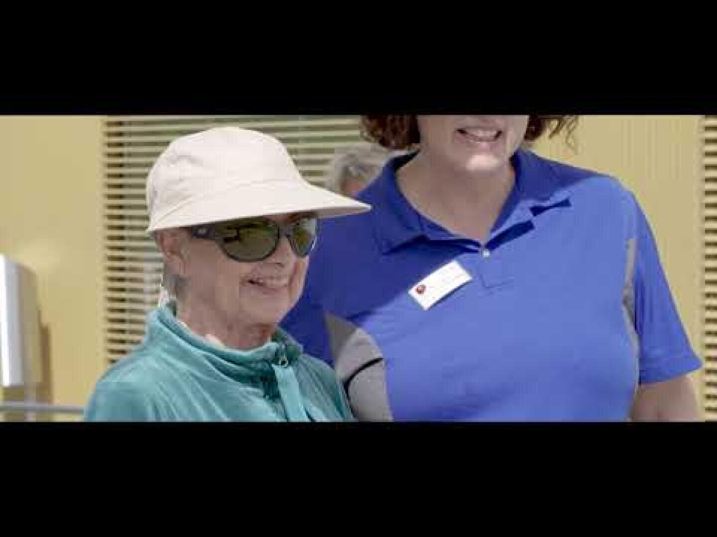 Deux femmes se tiennent à l'extérieur ; l'une d'elles porte un chapeau de soleil, des lunettes de soleil et une veste bleu sarcelle, et elle sourit. L'autre, dont on ne voit qu'une partie hors champ, porte une chemise bleue à col avec un badge nominatif.