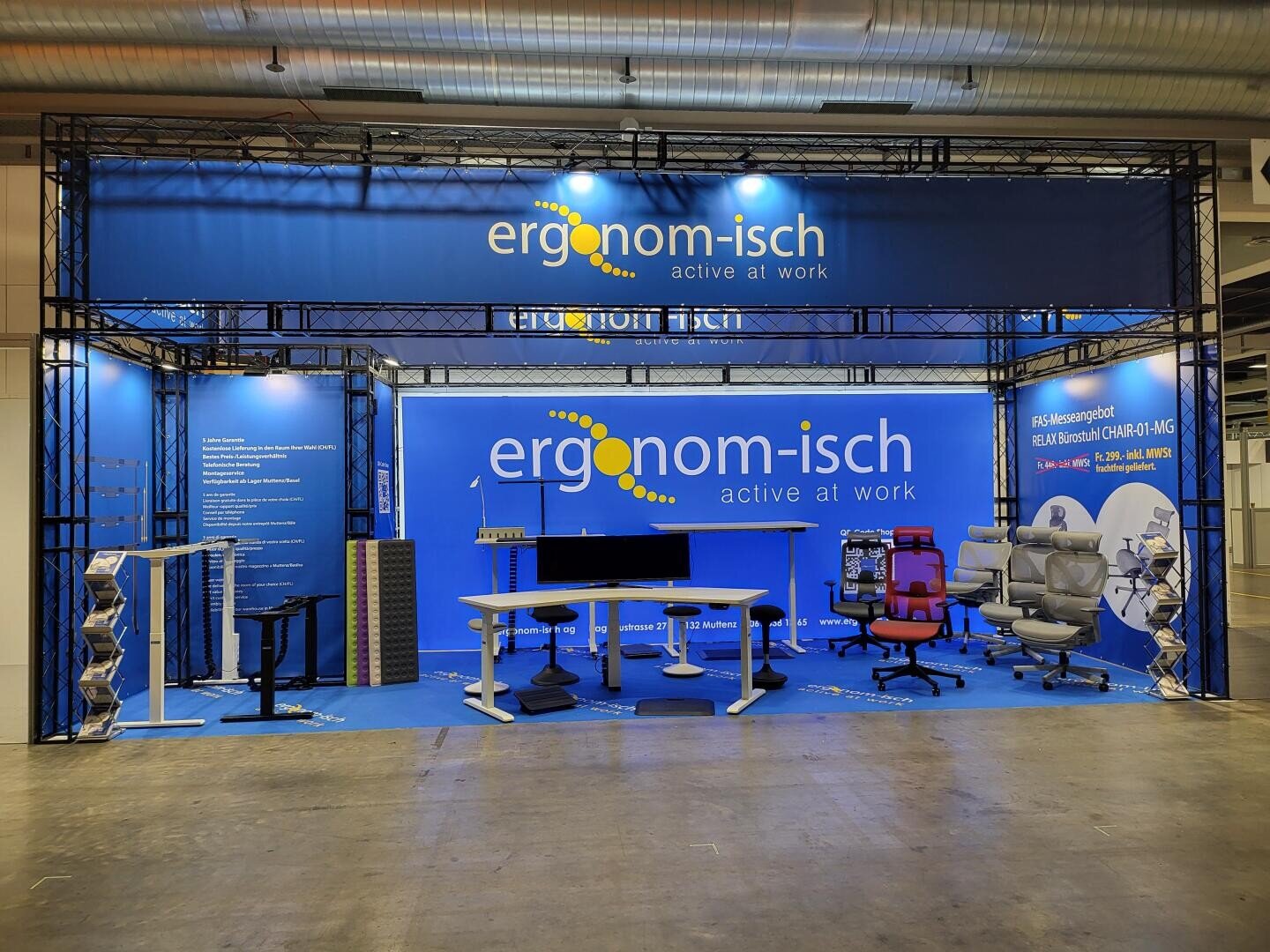 Stand d'exposition pour « ergonom-isch » présentant du mobilier de bureau ergonomique, notamment des chaises, des bureaux et des accessoires, avec une identité visuelle bleue et des bannières d'information. Le stand est bien éclairé et aménagé de manière claire pour les visiteurs.