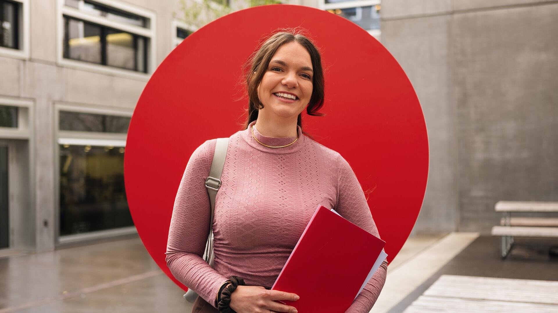 Une jeune femme, munie d'un sac à dos et d'une chemise rouge, sourit alors qu'elle se tient à l'extérieur devant un grand cercle rouge ; on aperçoit des bâtiments modernes en arrière-plan.