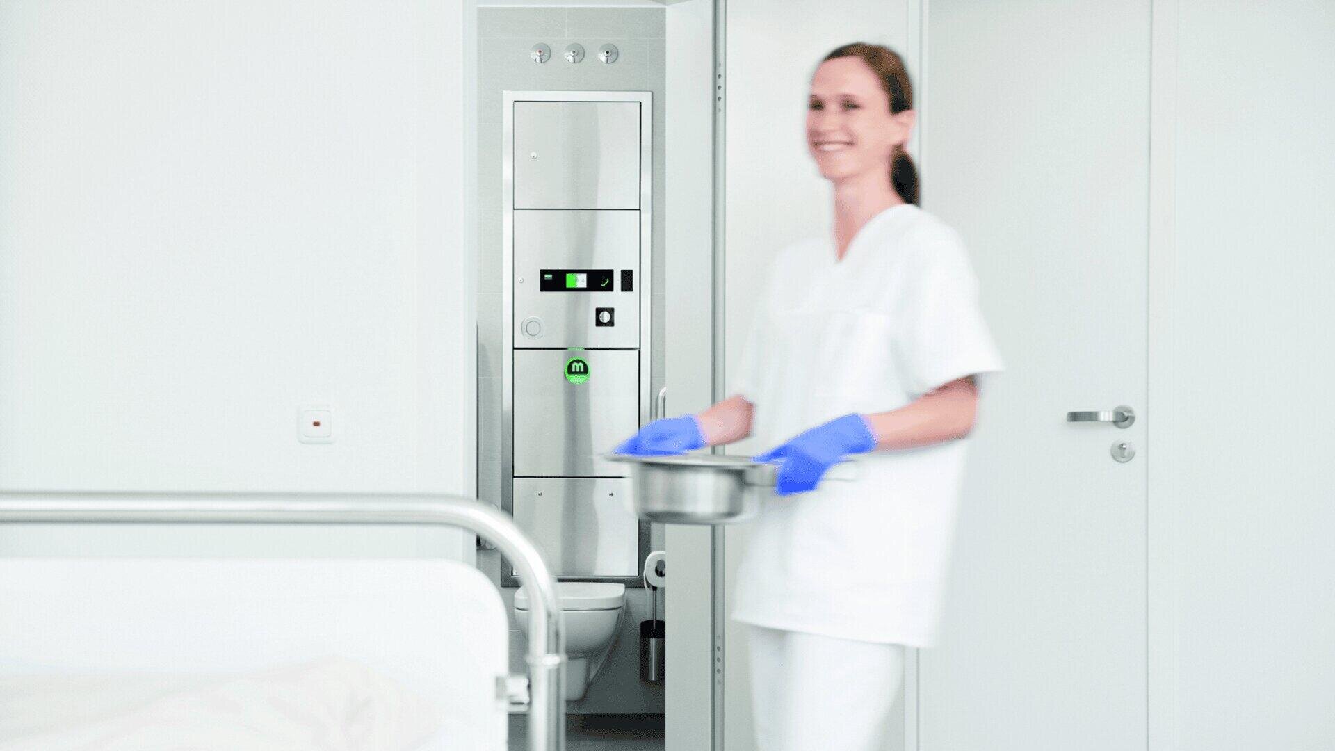 Eine Krankenschwester mit blauen Handschuhen hält einen Metallbehälter in der Hand, lächelt und geht durch ein Krankenzimmer mit weißen Wänden, in dessen Hintergrund ein Gerät der Marke MEIKO, Modell TopLine, in der Wand oberhalb der Toilettenschüssel eingebaut ist.
