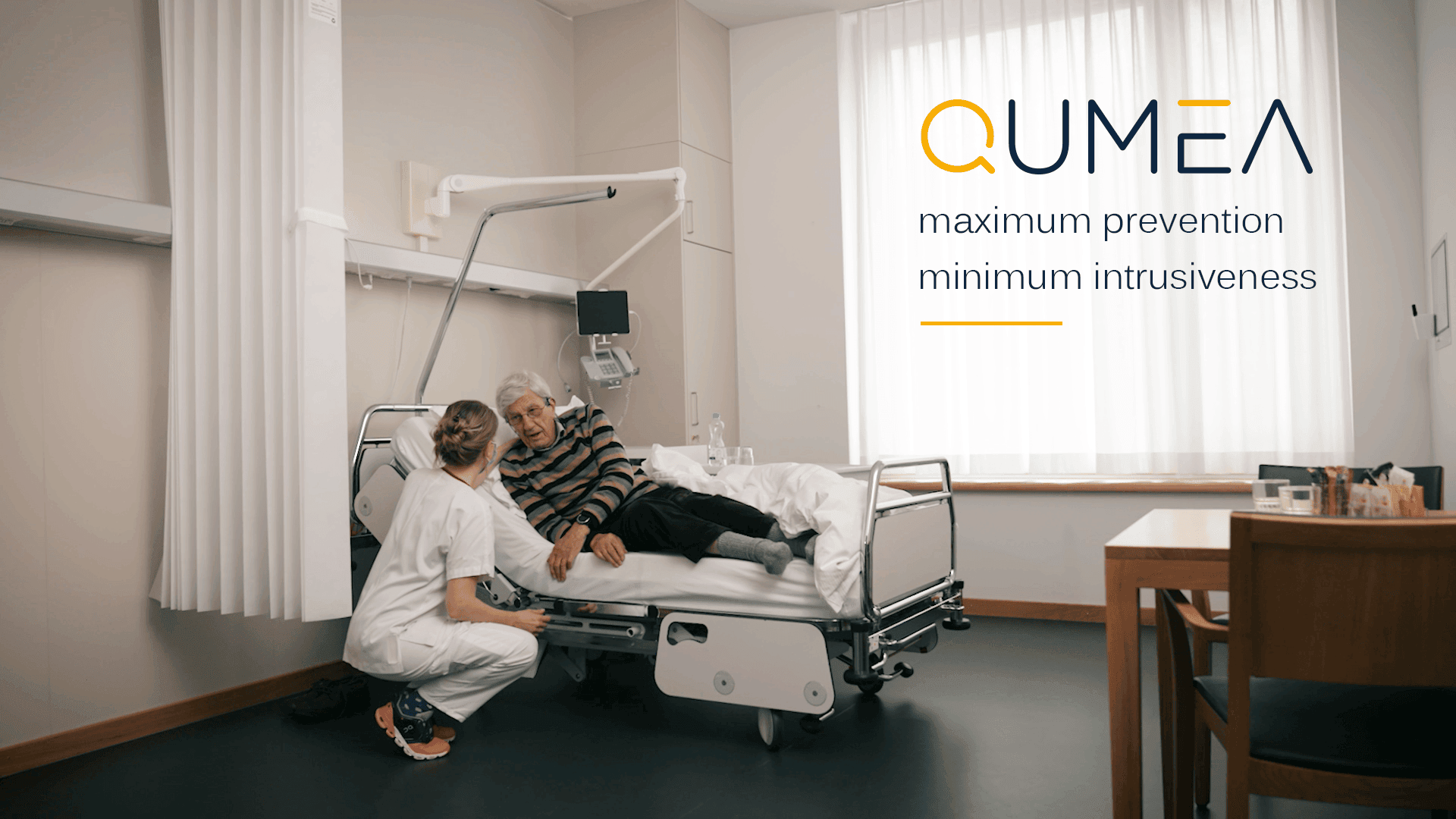 Eine Krankenschwester kniet neben einem älteren Patienten, der aufrecht in einem Krankenhausbett sitzt, und stützt ihn. Das Sonnenlicht fällt durch große Fenster. Der Text lautet: QUMEA - maximale Prävention, minimale Aufdringlichkeit.