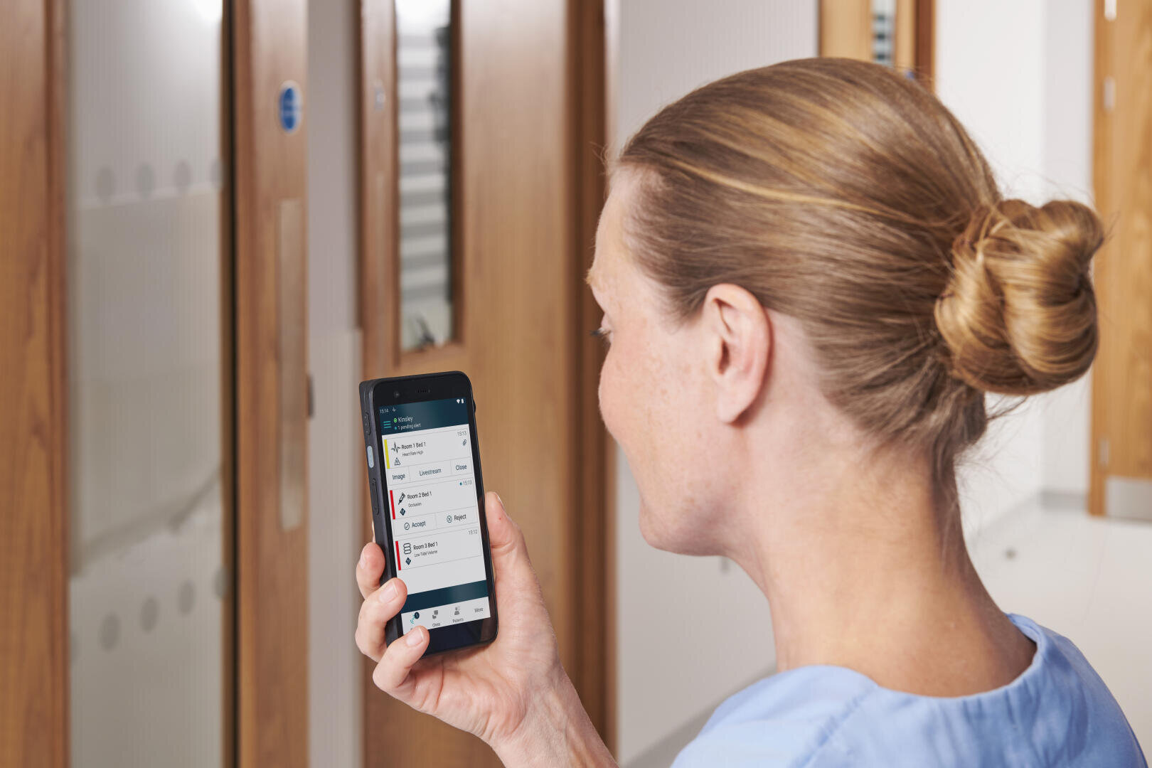 Eine Frau mit blonden Haaren, die zu einem Dutt zusammengebunden sind, schaut auf ein Smartphone, auf dem eine medizinische App angezeigt wird, während sie in einem Flur mit Holztüren steht.