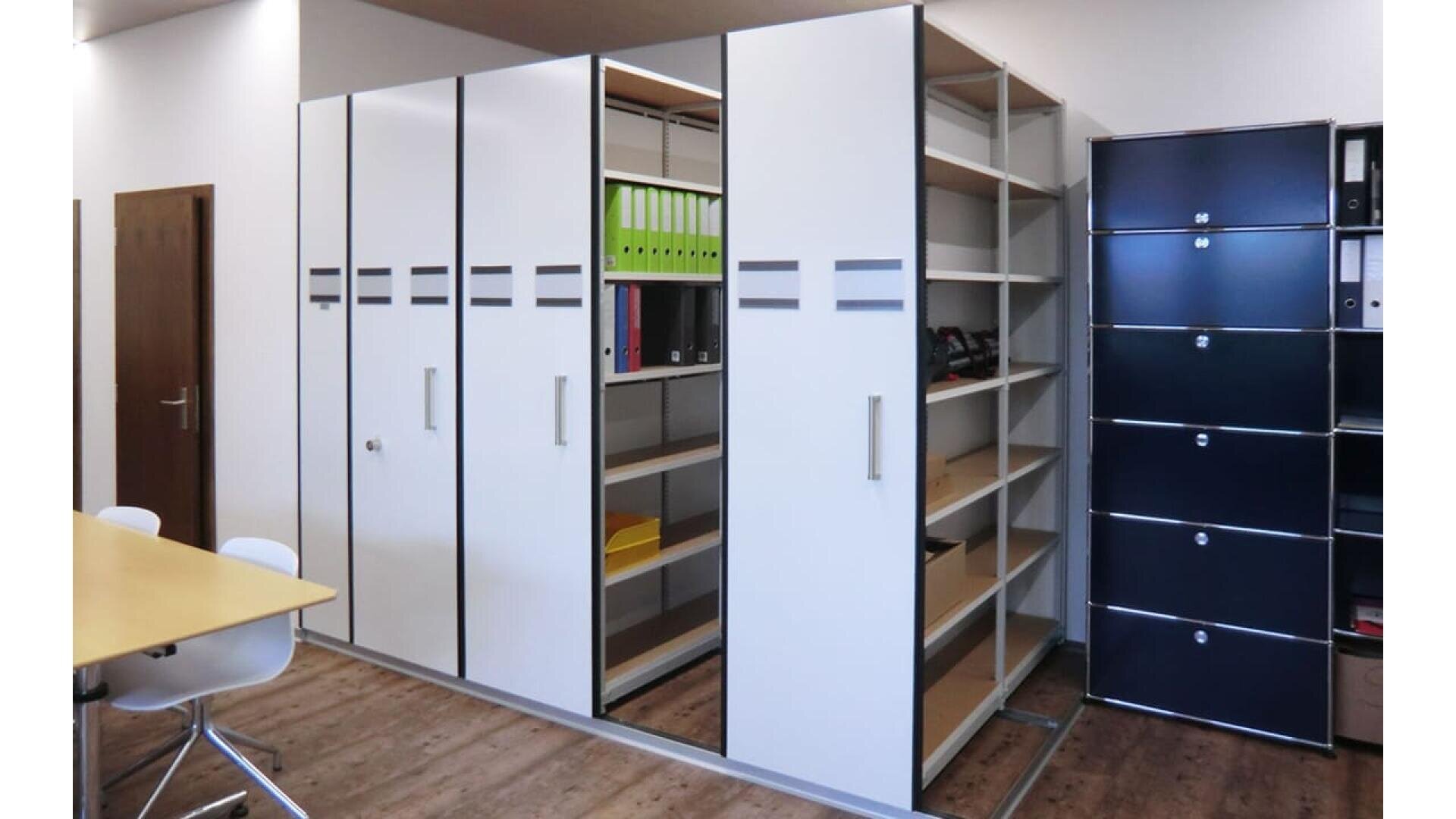 Un bureau moderne avec des armoires de rangement blanches coulissantes, dont certaines s'ouvrent pour révéler des étagères contenant des dossiers et des fournitures de bureau. À côté, des meubles de rangement noirs et une table de réunion en bois avec des chaises blanches.