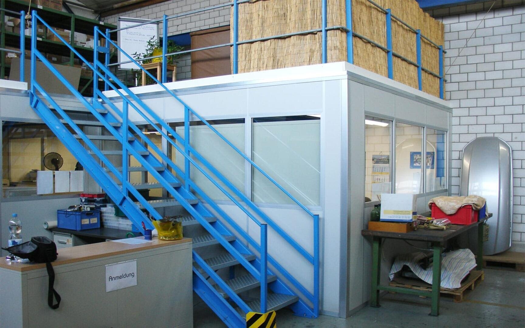 Ein moderner industrieller Arbeitsbereich mit einer blauen Metalltreppe, die zu einer oberen, mit Stroh bedeckten Plattform führt, darunter ein geschlossener Büroraum, Schreibtische, Geräte und verschiedene Objekte, die um den Bereich herum angeordnet sind.