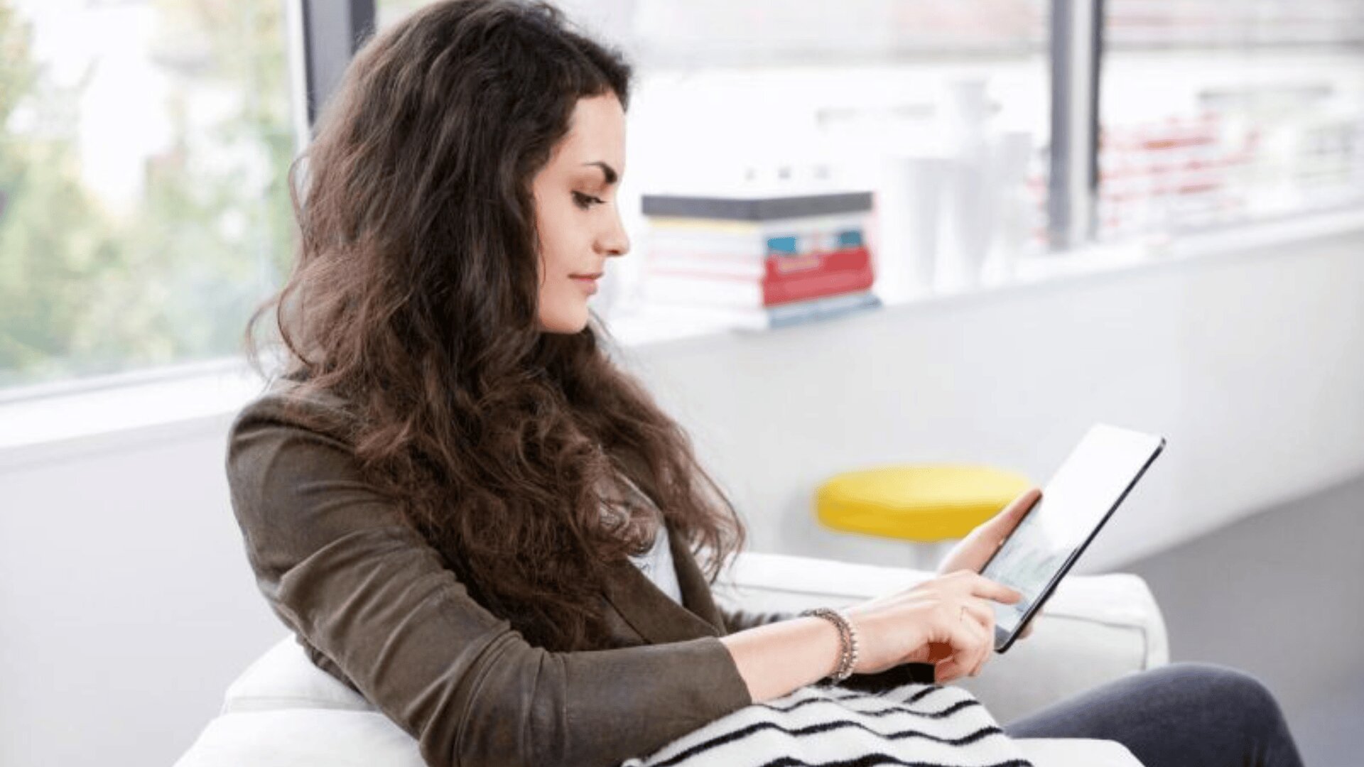 Eine Frau mit langen, lockigen braunen Haaren sitzt auf einer Couch in einem Haus und benutzt ein Tablet. Sie trägt einen grünen Blazer über einem hellen Hemd, und im Hintergrund stehen Bücher und ein gelber Hocker.