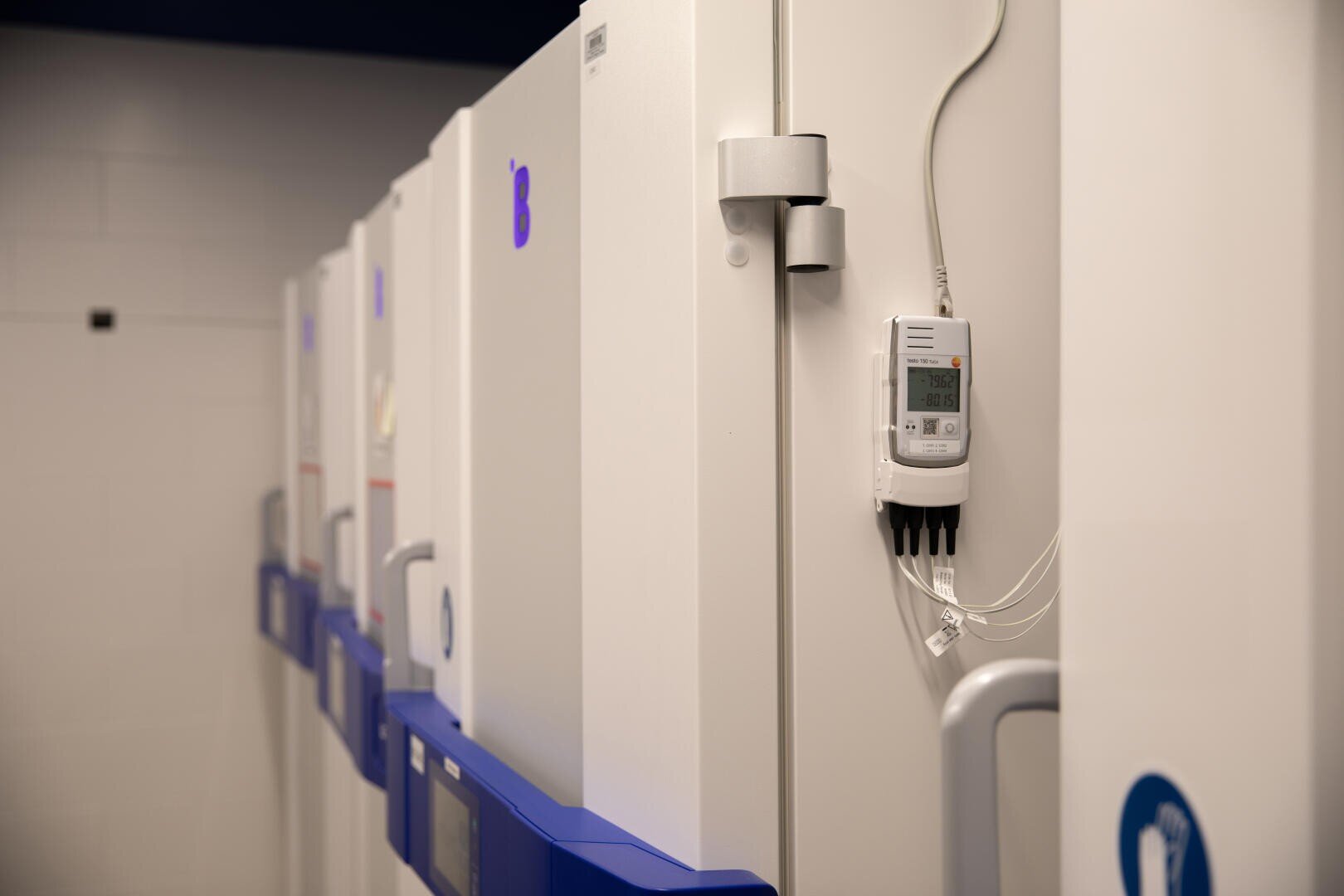 Une rangée de congélateurs industriels blancs dans un laboratoire. Un appareil de surveillance numérique fixé au congélateur le plus proche affiche des températures de -79,62 et -80,15 degrés Celsius. La lettre B est visible sur les étiquettes des portes.