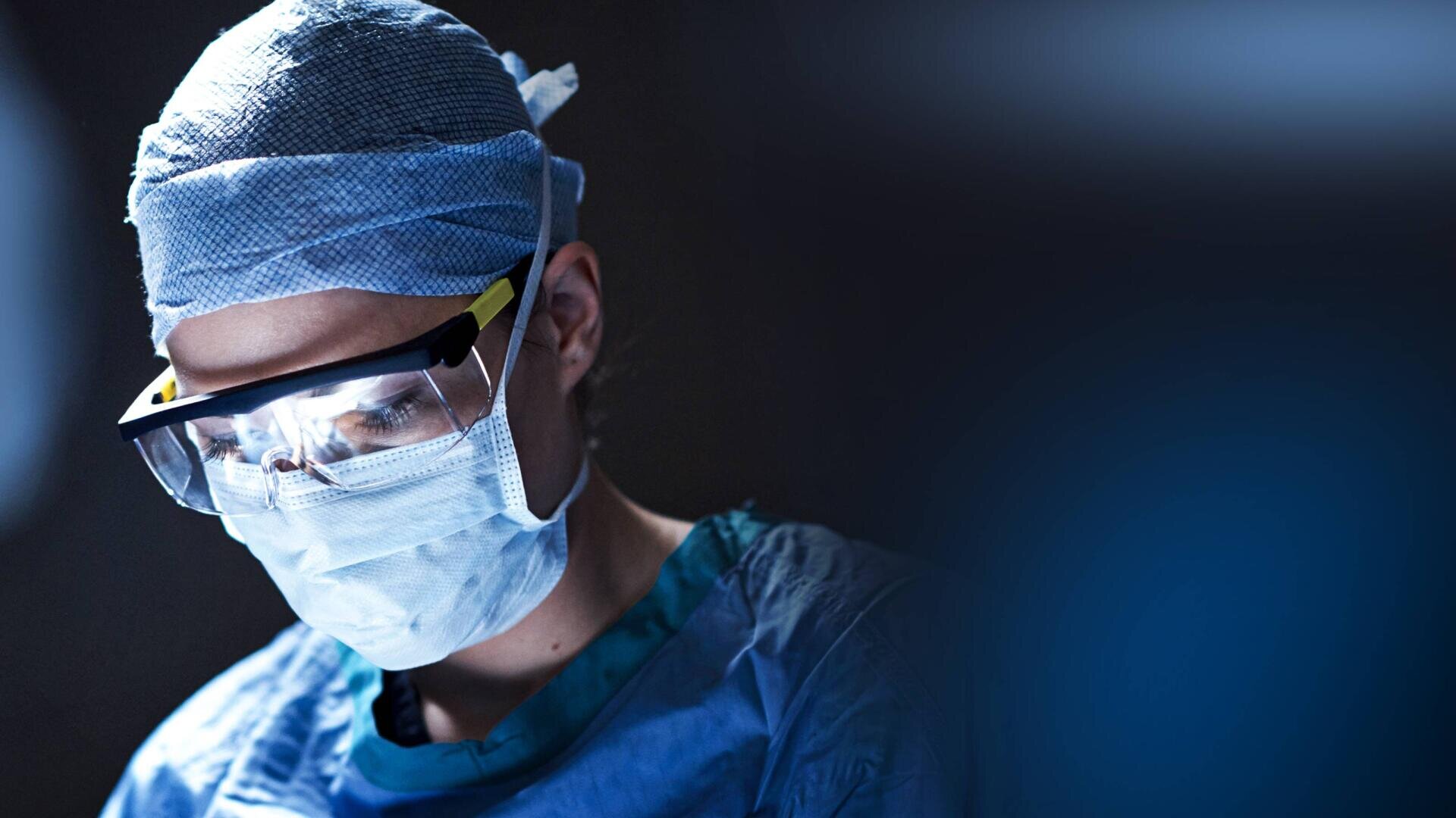 Ein Chirurg, der eine OP-Maske, eine OP-Haube und eine Schutzbrille trägt, ist während einer Operation voll konzentriert; er wird von einem hellen Licht vor einem dunklen, verschwommenen Hintergrund beleuchtet.
