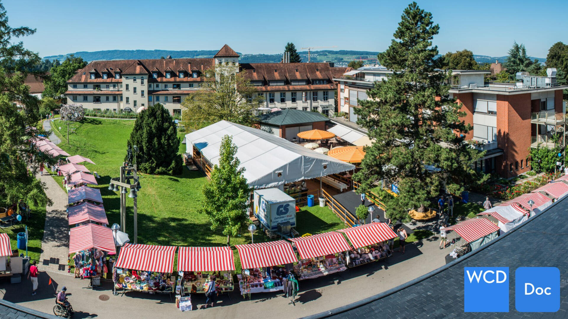 Ein lebhafter Markt im Freien mit rot-weiß gestreiften Ständen ist auf einem Weg neben großen Gebäuden und grünen Rasenflächen mit Bäumen und entfernten Hügeln unter einem klaren blauen Himmel aufgebaut. WCD- und Doc-Symbole erscheinen in der unteren rechten Ecke.