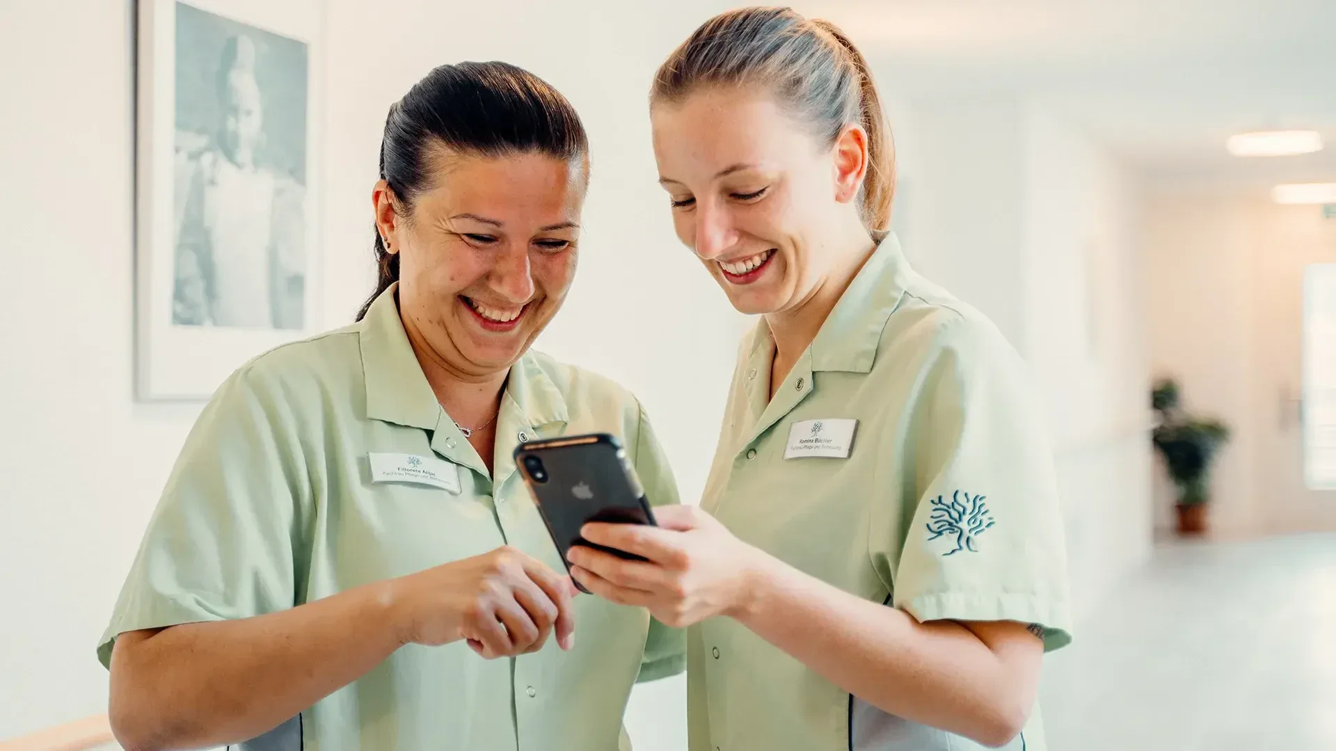 Deux professionnels de la santé en uniformes vert clair sourient et regardent ensemble un smartphone dans un couloir lumineux ; ils ont l'air heureux et engagés. Une photo encadrée est accrochée au mur derrière eux.