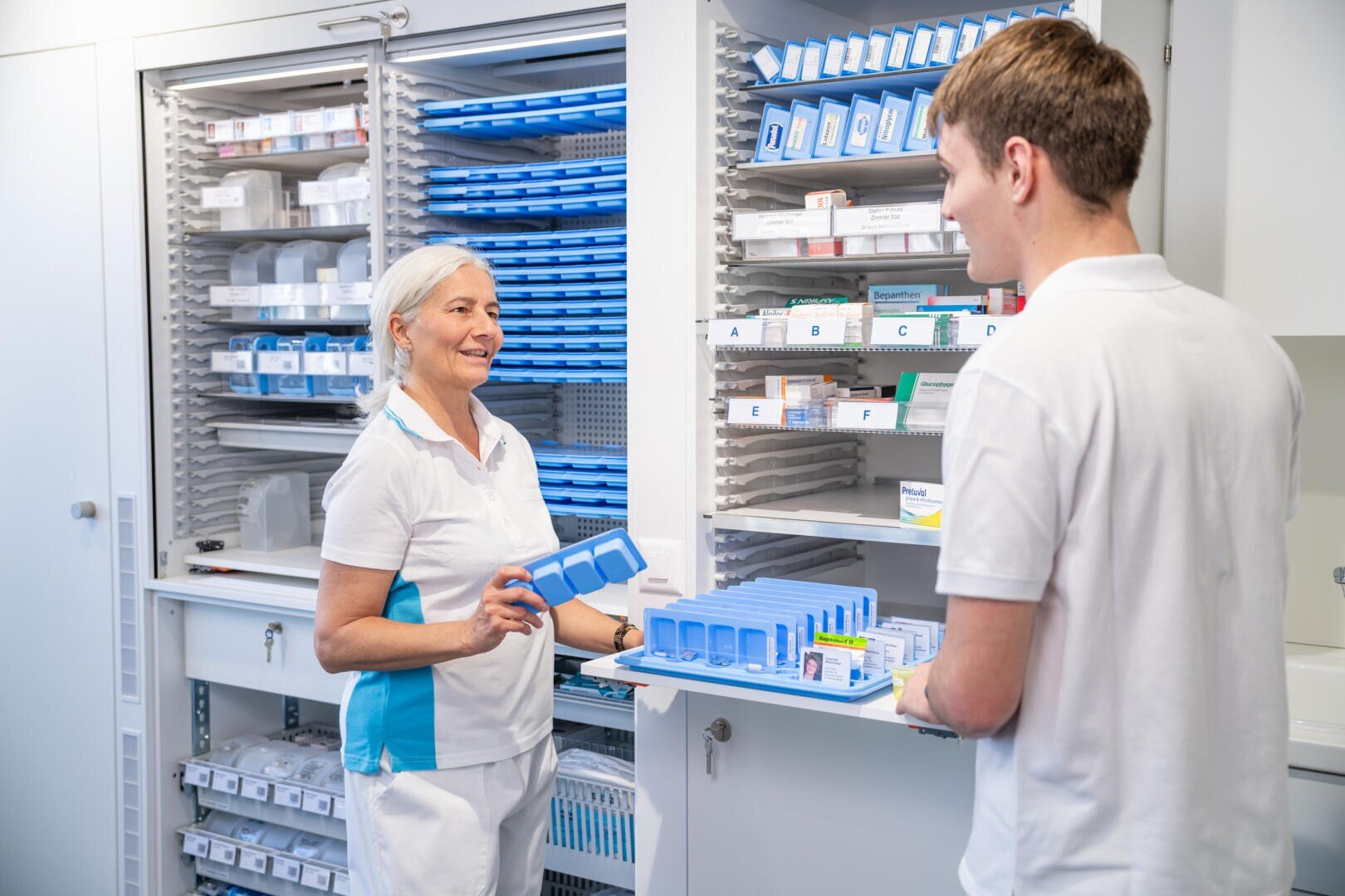 Zwei medizinische Fachkräfte in weißer Arbeitskleidung stehen in einer Apotheke; eine Frau hält einen blauen Tablettenbehälter in der Hand, während sie mit einem männlichen Kollegen spricht, der ein Tablett mit Medikamenten und blauen Tablettenbehältern in der Hand hält. Im Hintergrund sind Regale mit Medikamentenschachteln zu sehen.