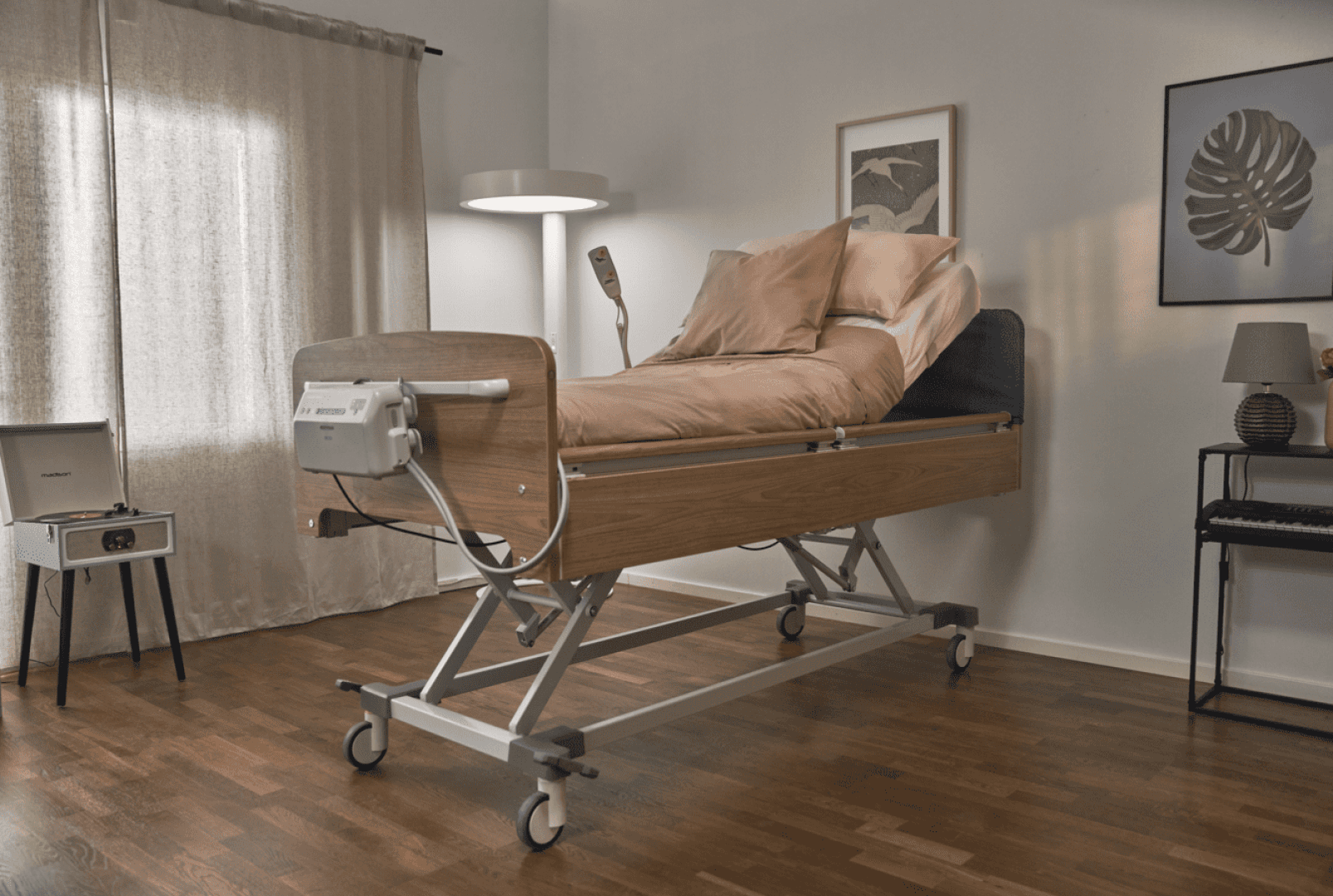 In einem gemütlichen, hell beleuchteten Zimmer mit Holzboden, einer Lampe, gerahmten Bildern, einem Fenster mit Vorhängen und einem Plattenspieler steht ein Krankenhausbett mit hellbrauner Bettwäsche.