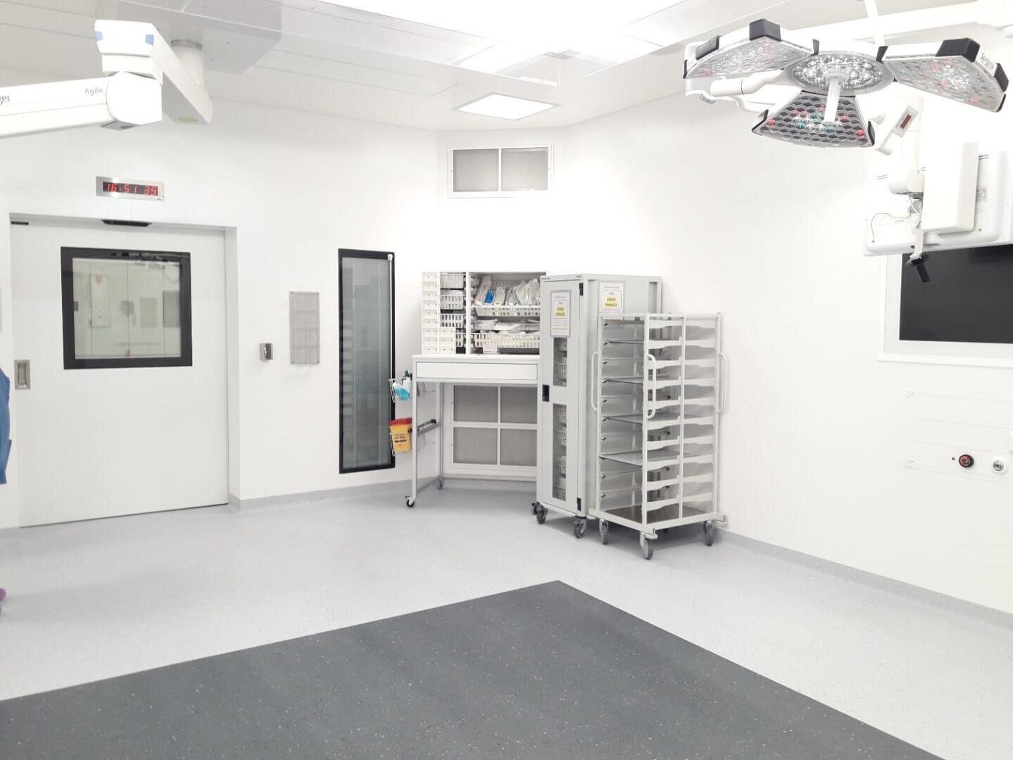 Une salle d'opération propre et bien éclairée avec des murs blancs, des équipements médicaux, des chariots de fournitures, des éclairages chirurgicaux suspendus et des portes à double battant. Le sol est gris clair avec un tapis plus foncé au centre.