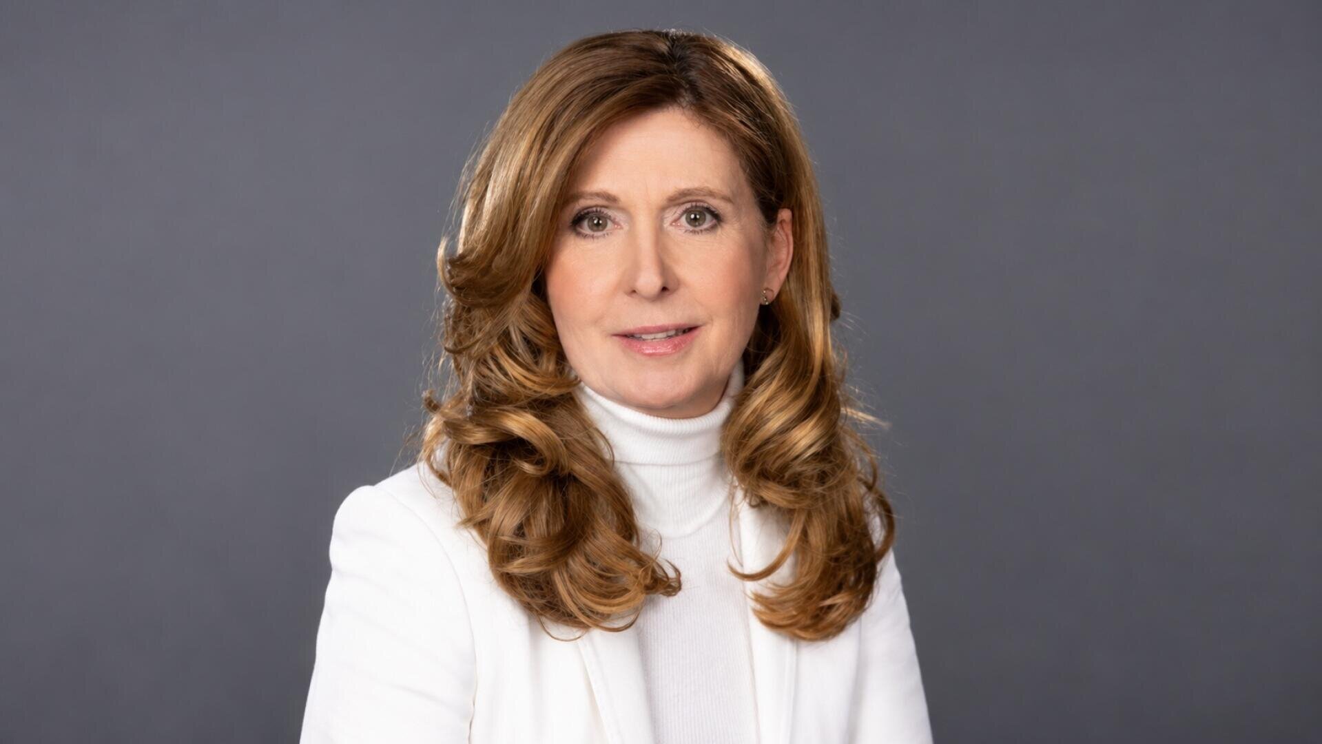 Eine Frau mit langen, gewellten, hellbraunen Haaren, die eine weiße Jacke und einen Rollkragenpullover trägt, posiert vor einem schlichten grauen Hintergrund und blickt mit ausdruckslosem Blick direkt in die Kamera.