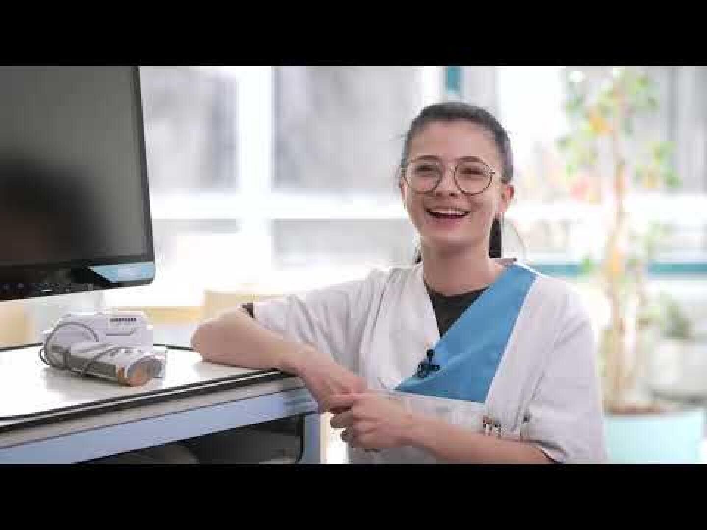Une femme souriante, vêtue d'une blouse chirurgicale et portant des lunettes, est assise dans une pièce lumineuse et moderne, à côté d'un écran d'ordinateur et d'appareils médicaux.