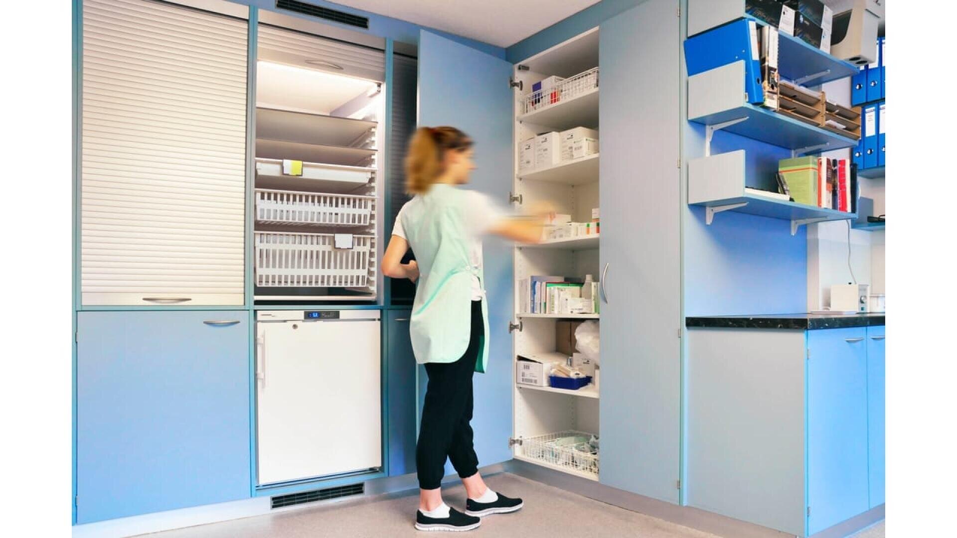 Une personne en blouse médicale se tient devant les armoires ouvertes d'une salle de fournitures médicales, cherchant à attraper les articles stockés sur les étagères. La pièce est équipée d'armoires bleues et de fournitures médicales organisées.