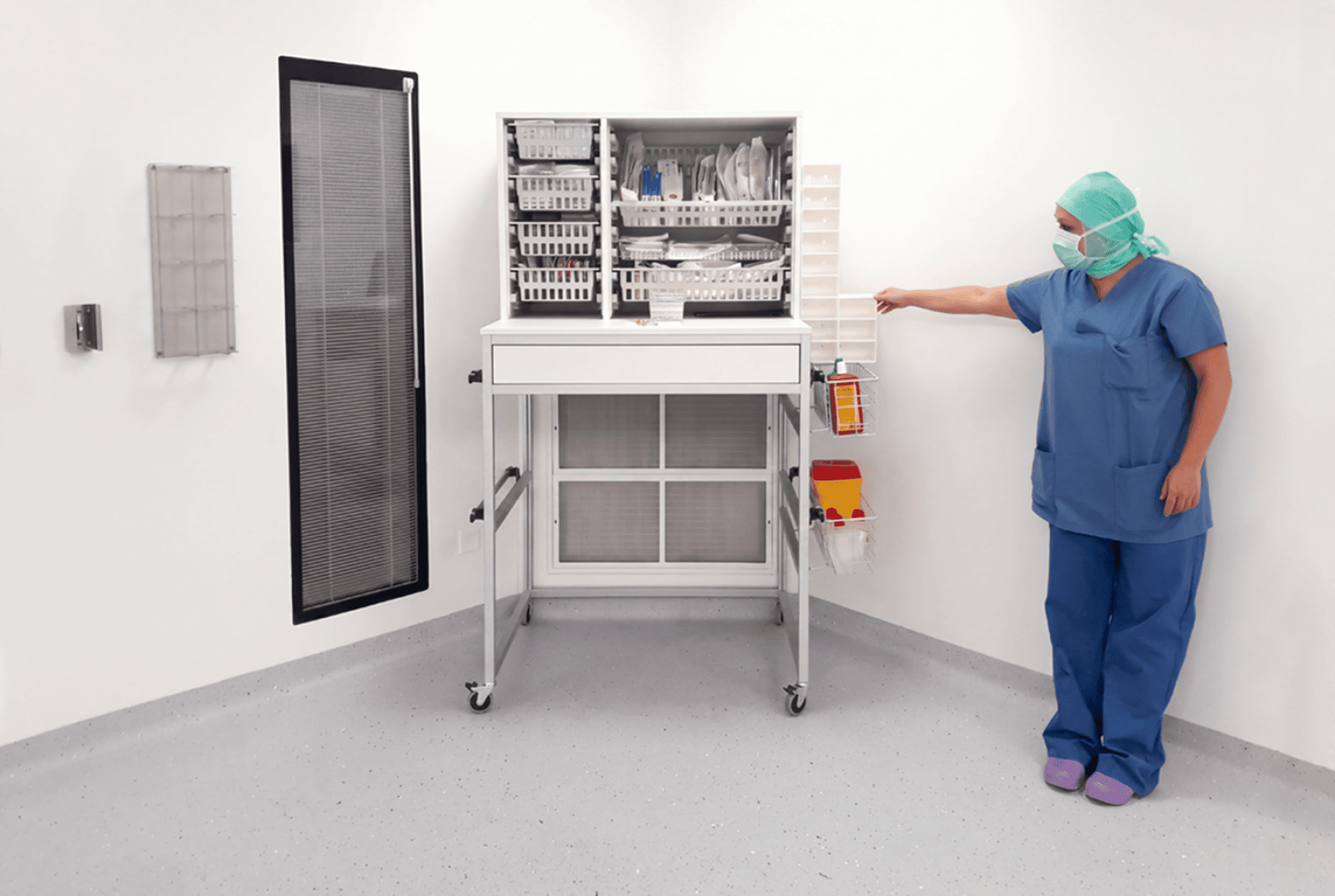 Ein Mitarbeiter des Gesundheitswesens in OP-Kleidung und mit Maske steht in einem sauberen, weißen Raum und zeigt auf eine an der Wand montierte Entsorgungseinheit neben einem hohen, offenen Schrank für medizinische Verbrauchsmaterialien.