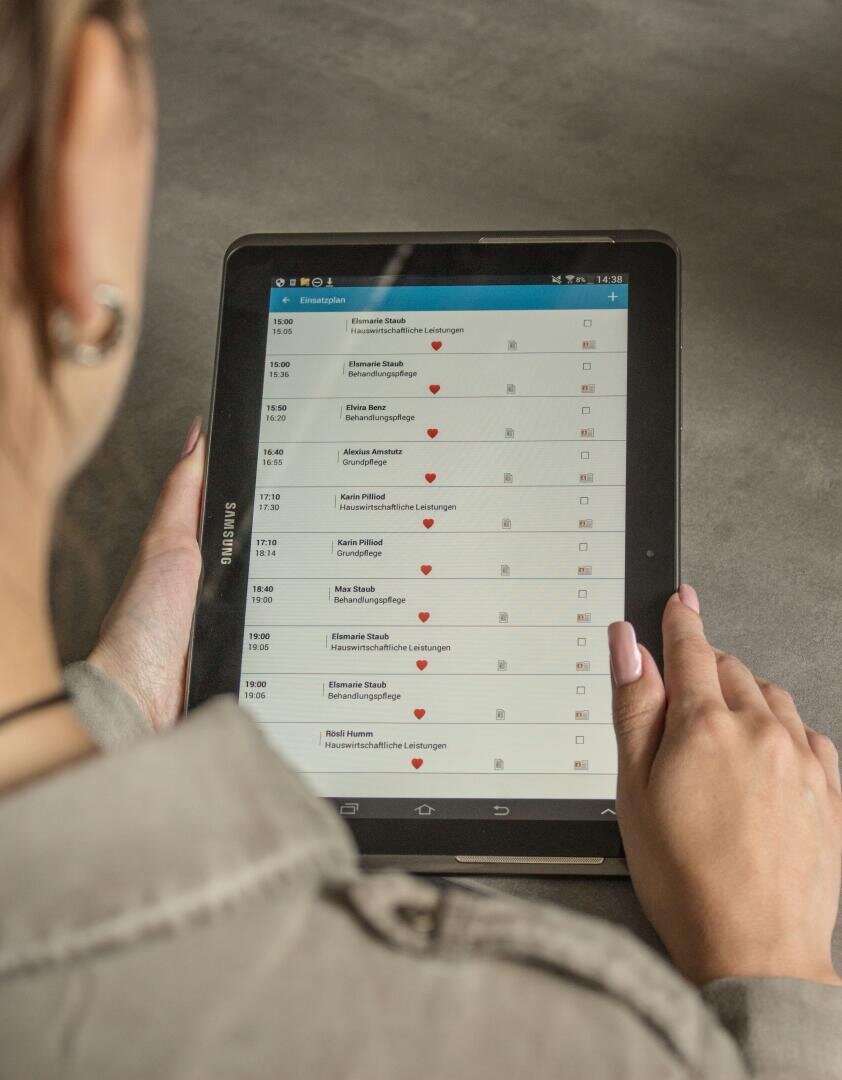 Eine Person mit langen Haaren und einem grauen Hemd hält ein Tablet in der Hand, auf dem ein digitaler Terminkalender oder eine Kontaktliste mit Namen, Uhrzeiten und roten Herzsymbolen neben einigen Einträgen angezeigt wird. Das Tablet ist von der Marke Samsung.