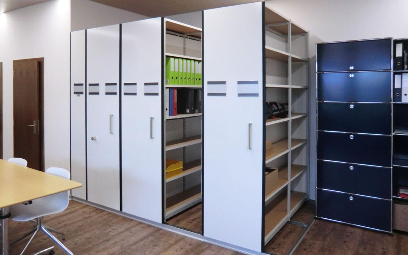 Weiße, verschiebbare Aktenschränke und offene Regale, gefüllt mit Ordnern und Büromaterial, stehen an einer Wand in einem modernen Büro mit Holzfußboden. Ein Tisch und Stühle sind teilweise im Vordergrund zu sehen.