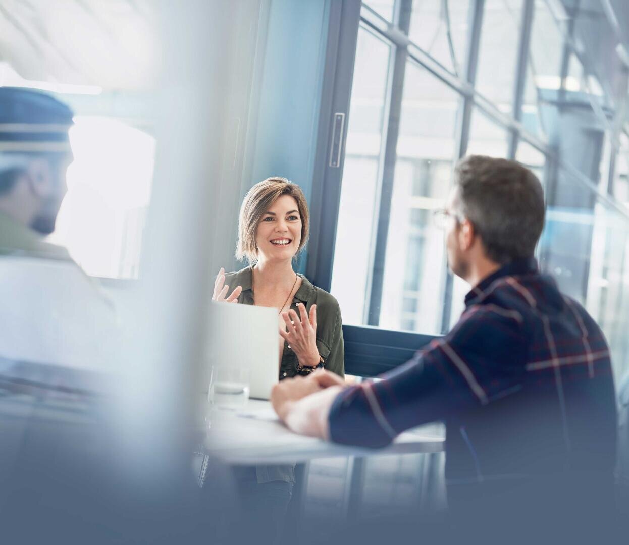 Drei Personen sitzen an einem runden Tisch in einem modernen Büro und unterhalten sich. Eine Frau in der Mitte lächelt, während sie spricht, und zwei Männer hören aufmerksam zu. Durch die großen Fenster strömt Tageslicht herein.