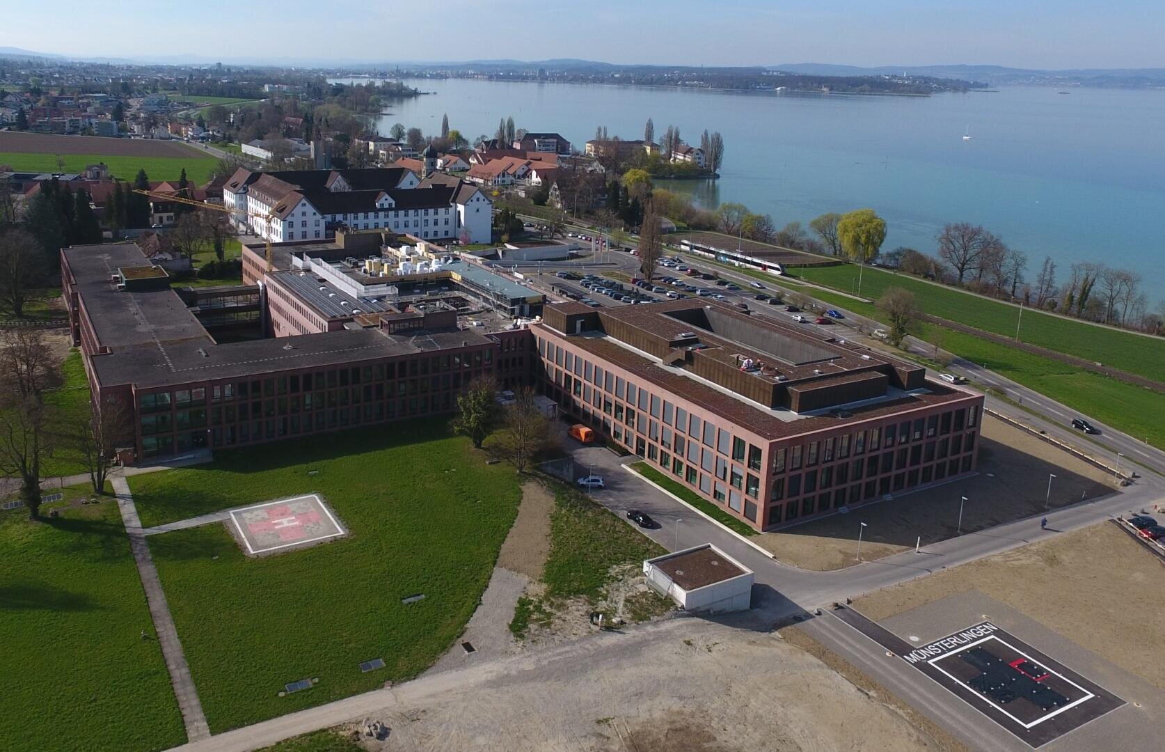 Luftaufnahme eines großen Krankenhauskomplexes in der Nähe eines Sees, mit mehreren Gebäuden, Parkplätzen, grünen Rasenflächen und zwei Hubschrauberlandeplätzen. In der Umgebung sind Häuser, Bäume und Wasser im Hintergrund zu sehen.