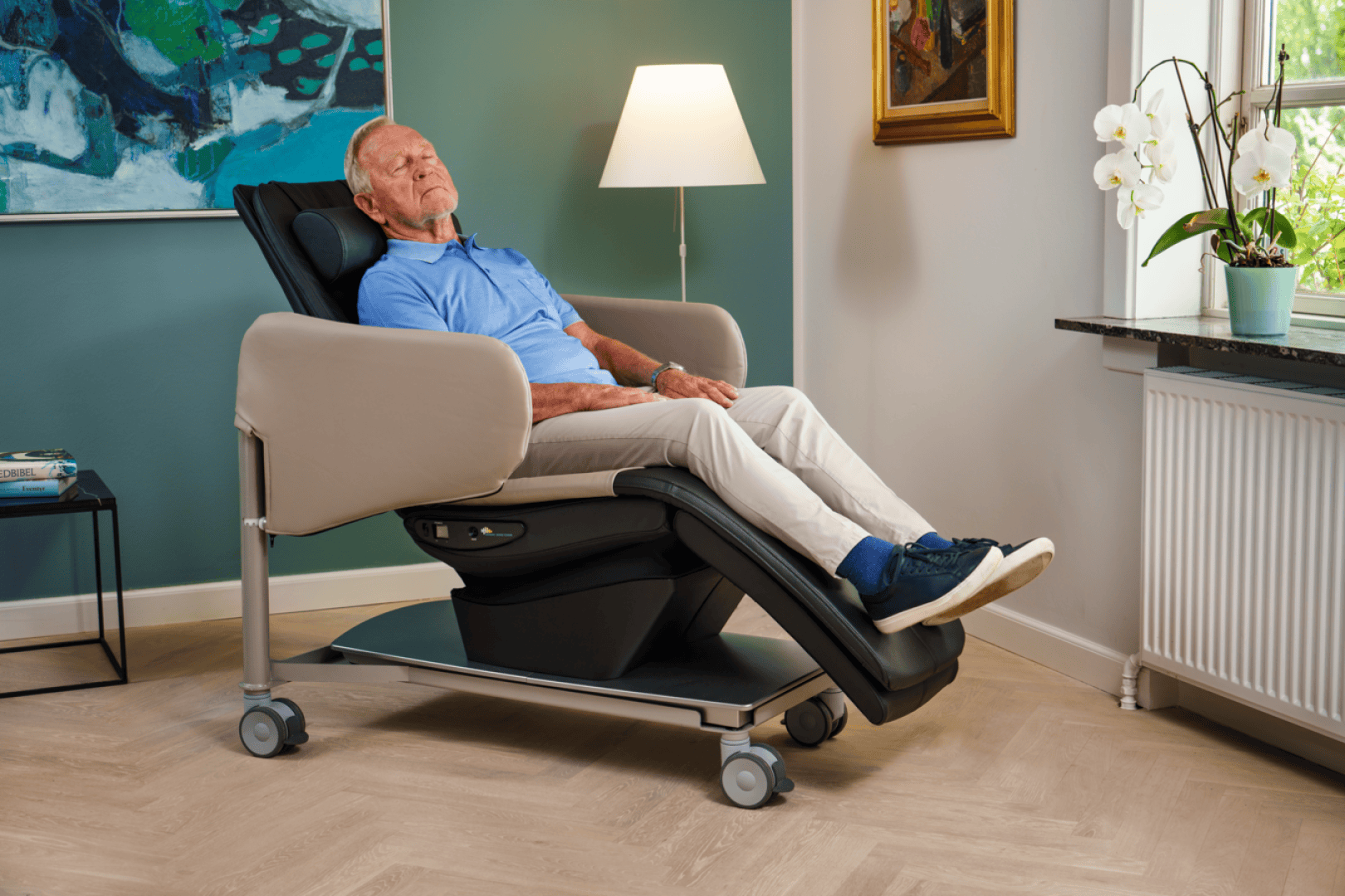 Un homme d'un certain âge, confortablement installé dans un fauteuil inclinable moderne à roulettes, dans un salon chaleureux et lumineux, près d'une fenêtre ornée de fleurs.