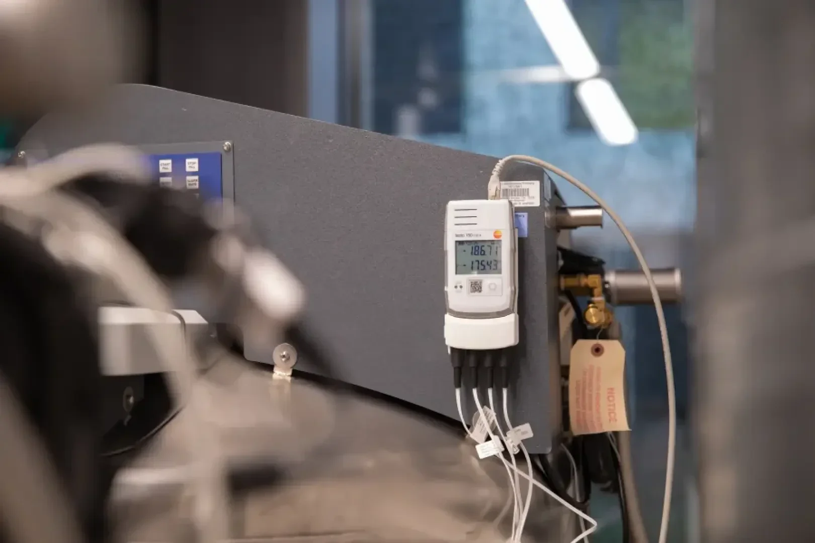 Dans une installation industrielle, un thermomètre numérique relié par des câbles affiche des valeurs de température. Un panneau est accroché à proximité, et on aperçoit au premier plan des pièces de machine floues.