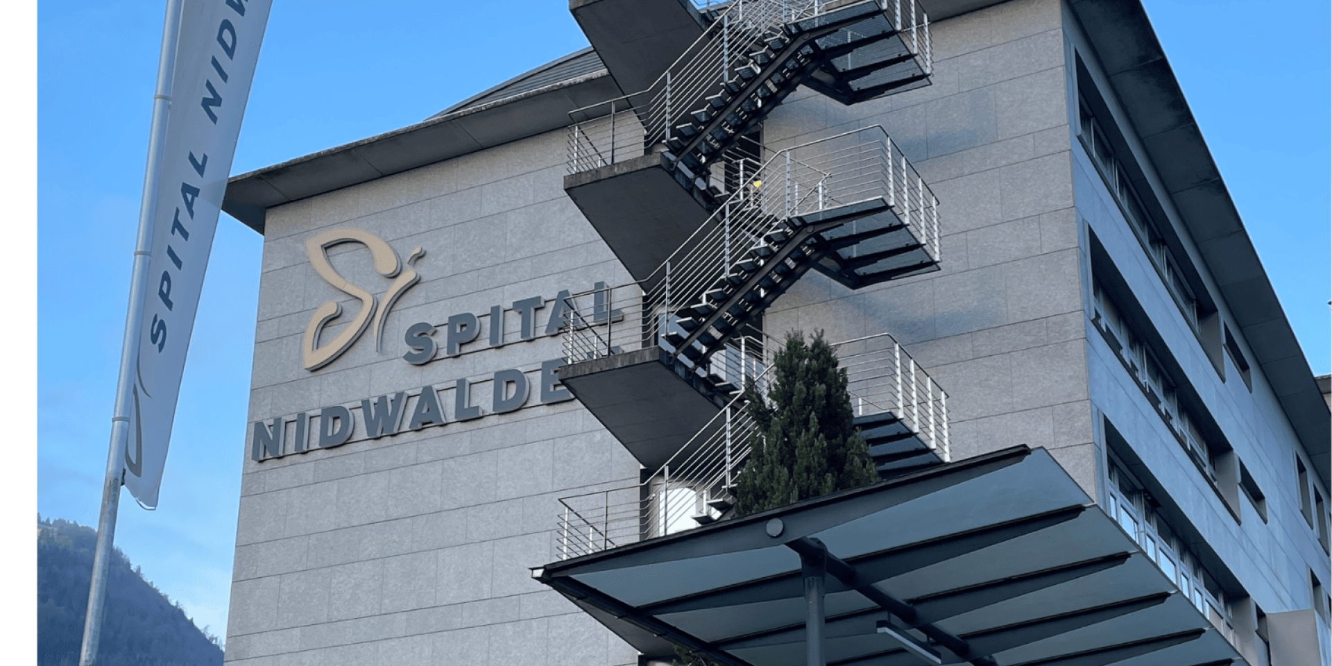 Die Außenansicht des Spitals Nidwalden, eines Krankenhausgebäudes mit einer modernen Feuerleiter aus Metall und einem Glasvordach. Im Vordergrund ist eine Fahne mit dem Namen des Spitals zu sehen.