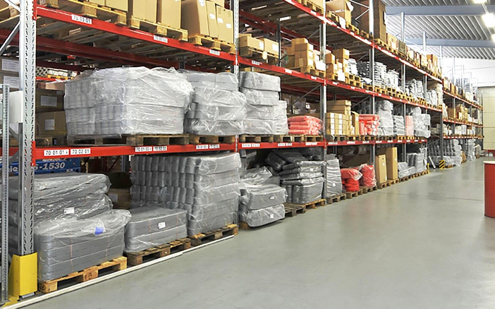 Großes Lager mit hohen Metallregalen, in denen sich Kisten und in Plastik verpackte Artikel stapeln, die auf Holzpaletten angeordnet sind. Der Boden ist sauber und geräumig, und der Bereich ist mit Oberlichtern gut beleuchtet.
