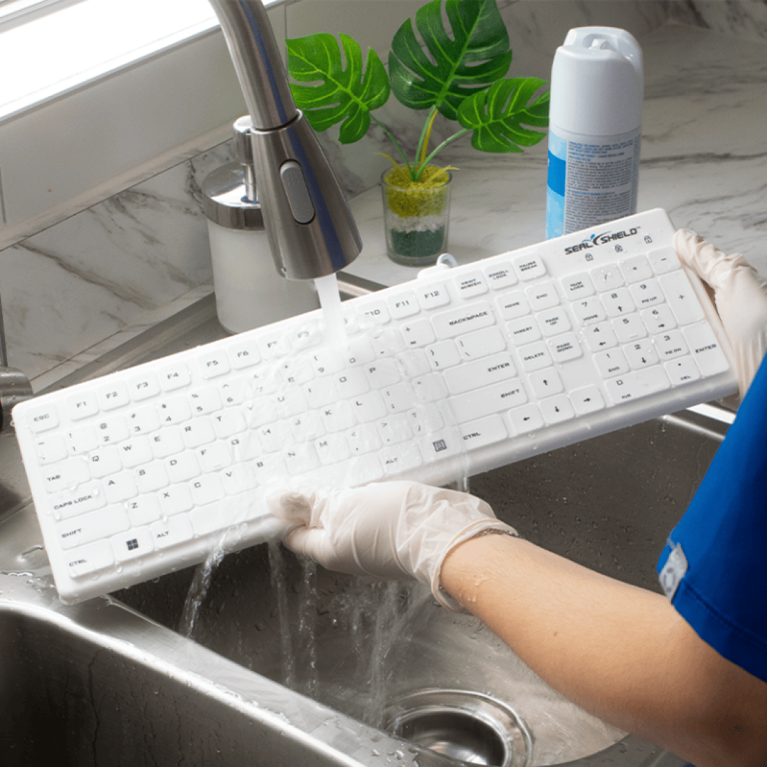 Une personne portant des gants rince un clavier d'ordinateur blanc sous l'eau courante dans l'évier. À l'arrière-plan, on aperçoit un flacon de produit nettoyant et une petite plante.