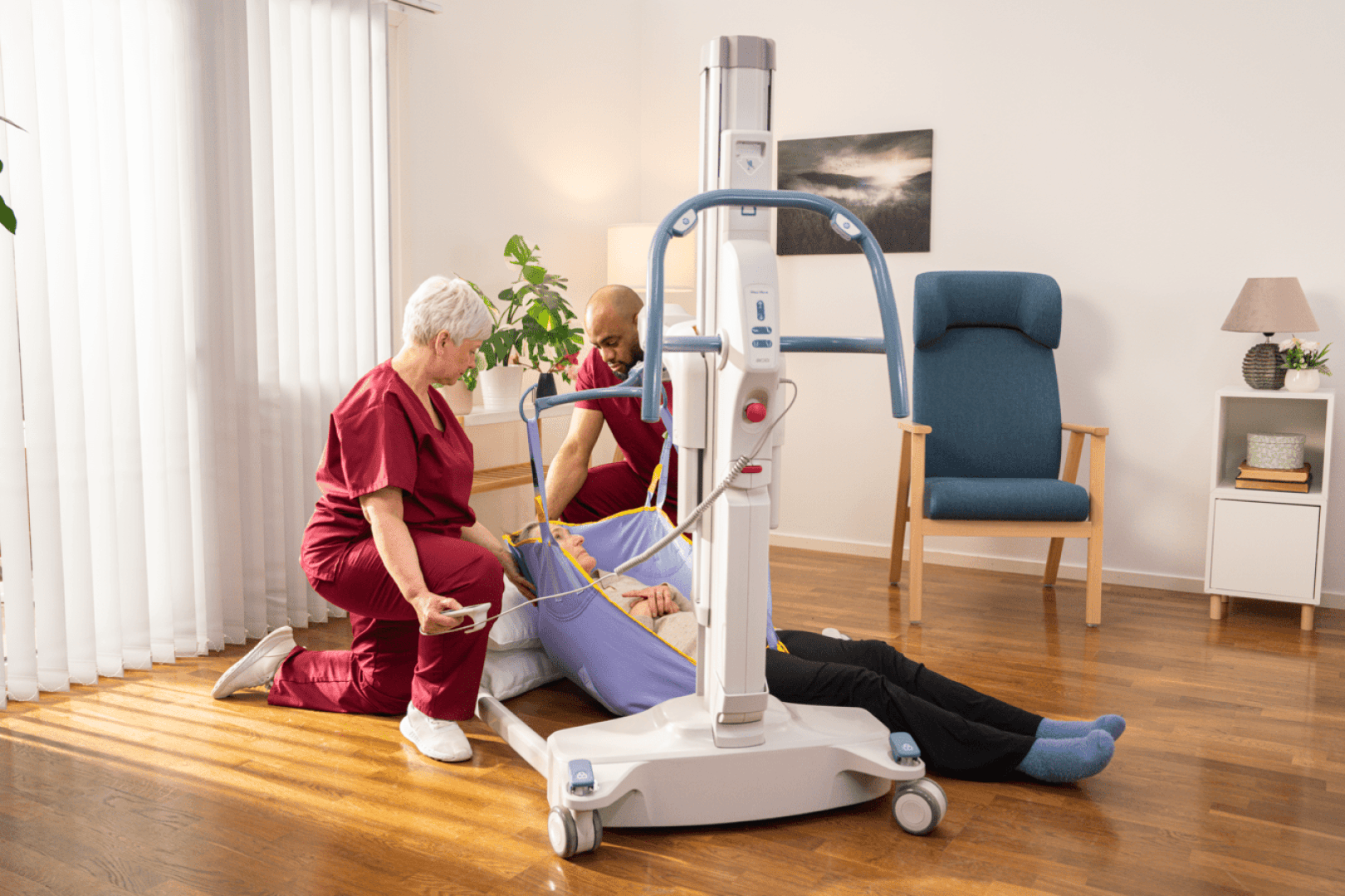 Deux professionnels de santé utilisent un lève-personne pour venir en aide à une personne allongée sur le sol, dans une pièce bien éclairée dotée d'un parquet, d'équipements médicaux et d'une chaise à l'arrière-plan.