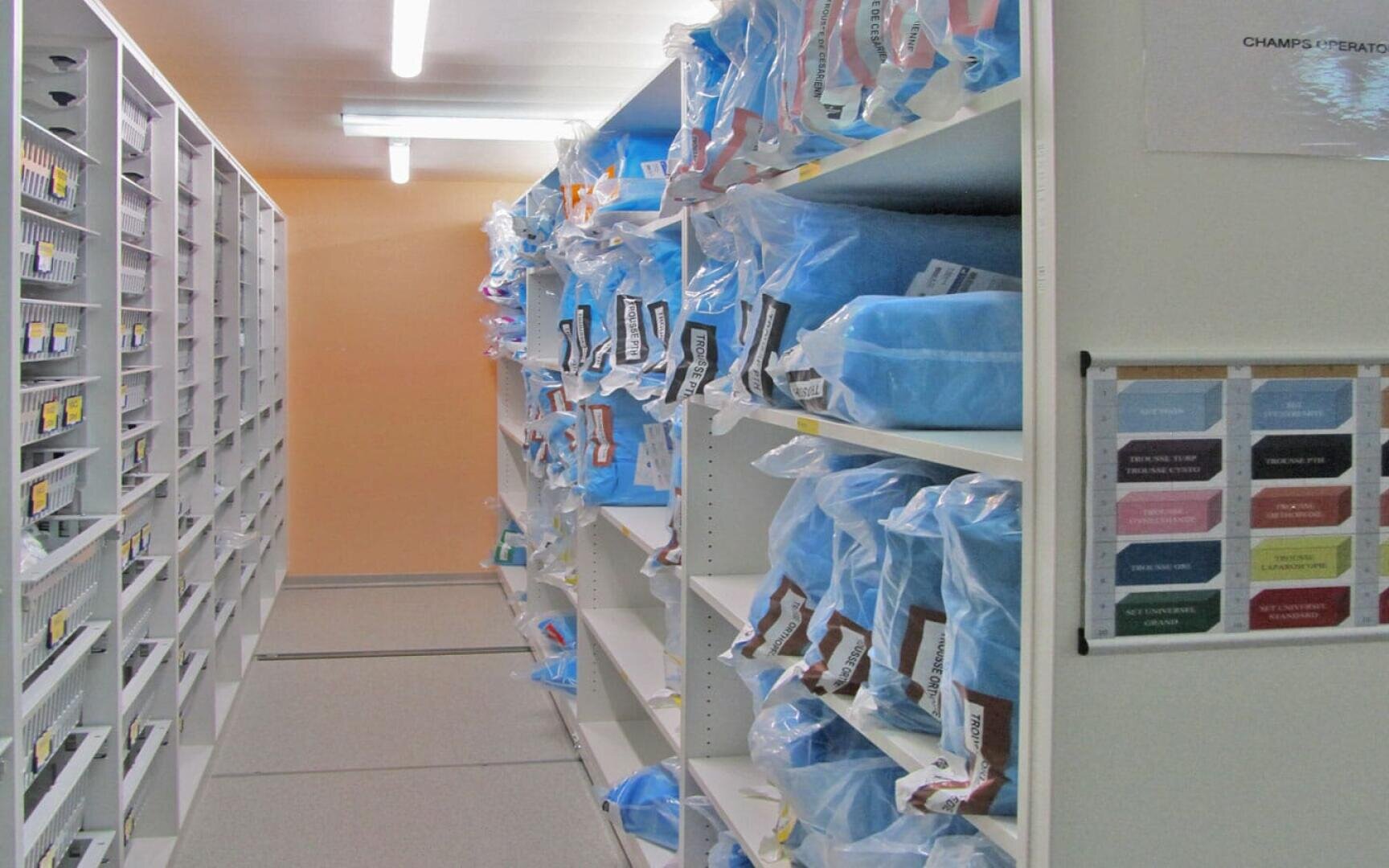 In einem hell erleuchteten Lagerraum sind die Regale mit blauen Tüten und beschrifteten Etiketten ausgekleidet, und an der Wand sind weitere Regale mit geordnetem Material und Tabellen zu sehen.
