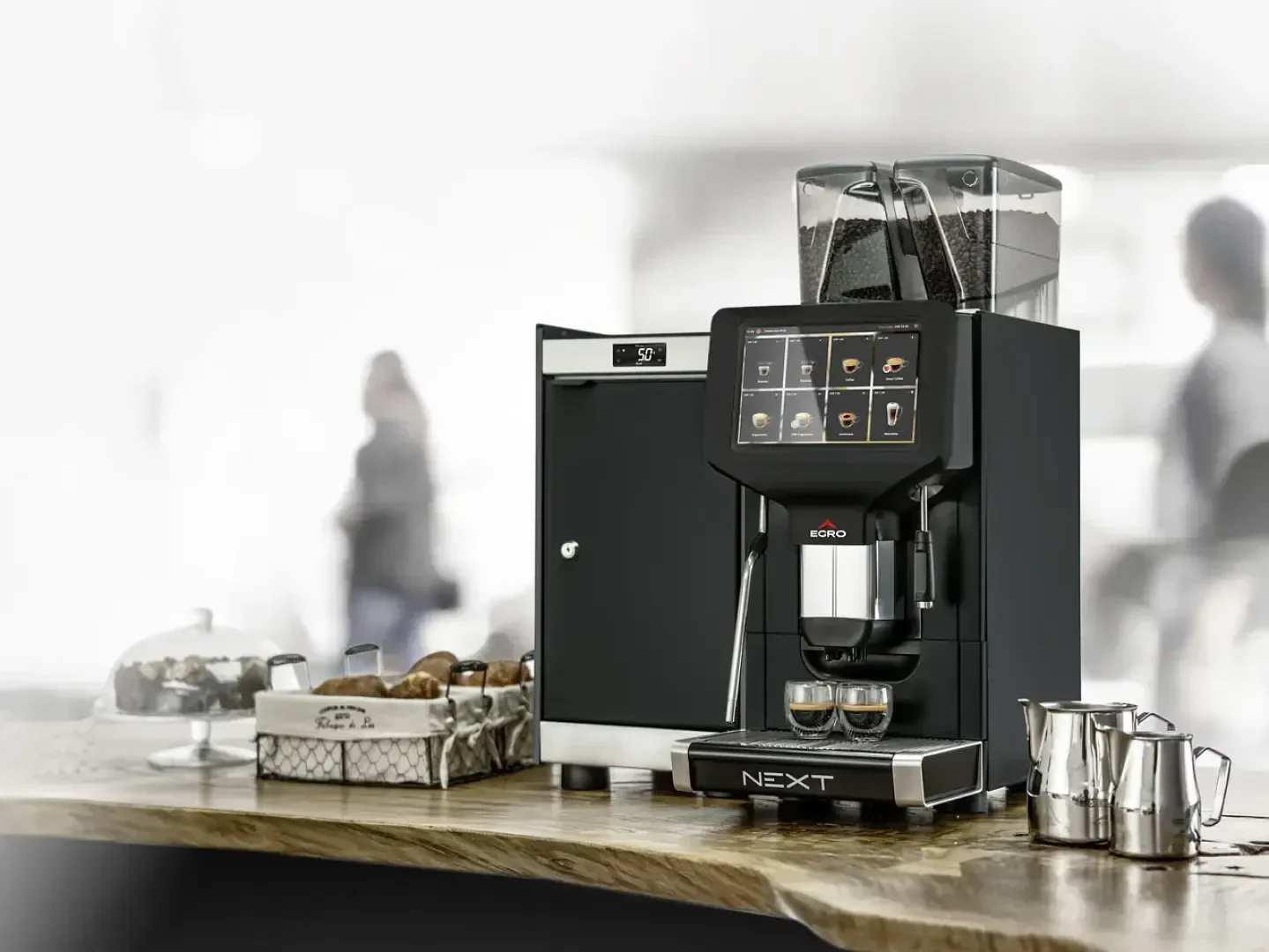 Sur un comptoir en bois se trouve une machine à café moderne dotée d'un écran tactile numérique, entourée d'un petit réfrigérateur, de tasses, d'un panier de viennoiseries et de pot à lait ; à l'arrière-plan, on aperçoit des silhouettes floues.