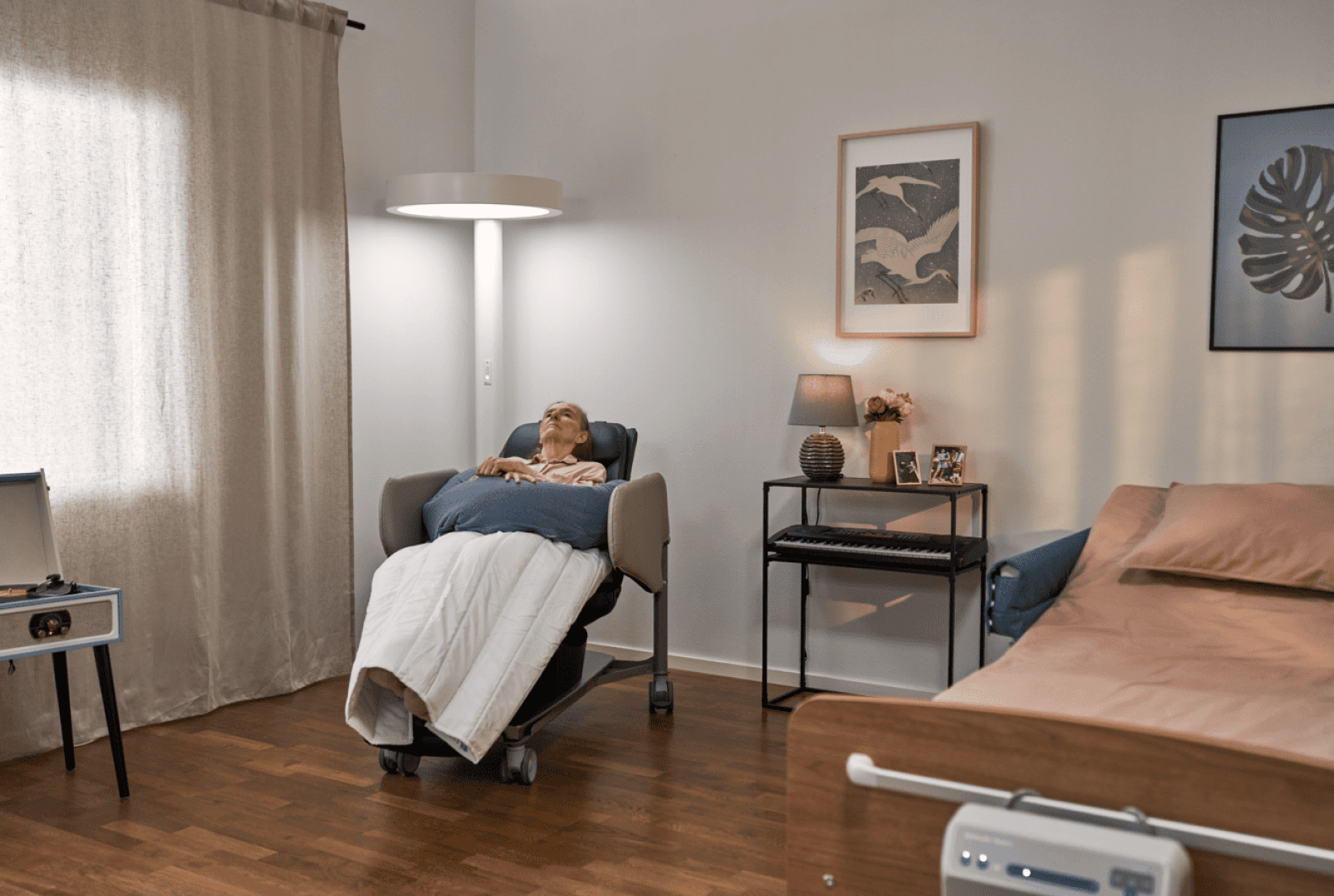 Une personne âgée se repose dans un fauteuil inclinable, recouverte d'une couverture, dans une chambre à coucher confortable et faiblement éclairée. La pièce est meublée d'un lit, d'une table de chevet, d'une lampe, de photos et de tableaux, ce qui crée une atmosphère calme et chaleureuse.