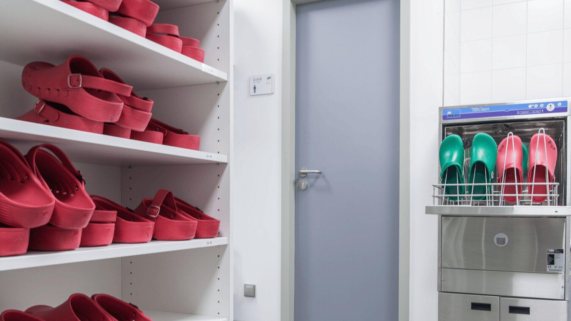 Regale mit roten Clogs, eine geschlossene graue Tür und ein Spülgerät der Marke MEIKO, Modell TopClean 60, in der rote und grüne Clogs liegen, in einem sauberen, gefliesten Raum in einem Krankenhaus.