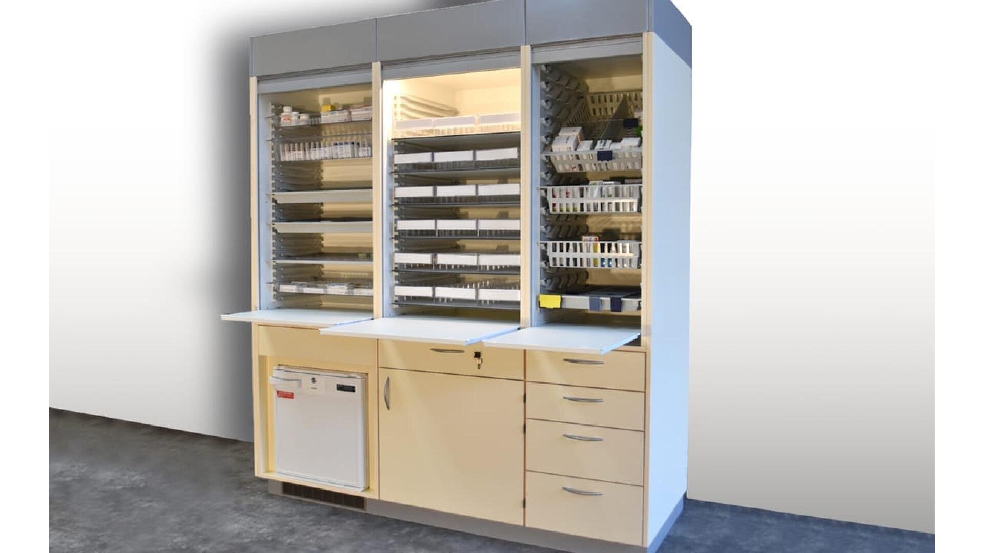 Une armoire de stockage médical avec des portes en verre présente des étagères organisées de plateaux et de conteneurs. En dessous, il y a des tiroirs, une petite armoire et un lave-vaisselle ou un stérilisateur compact intégré. L'armoire est posée sur un sol sombre.
