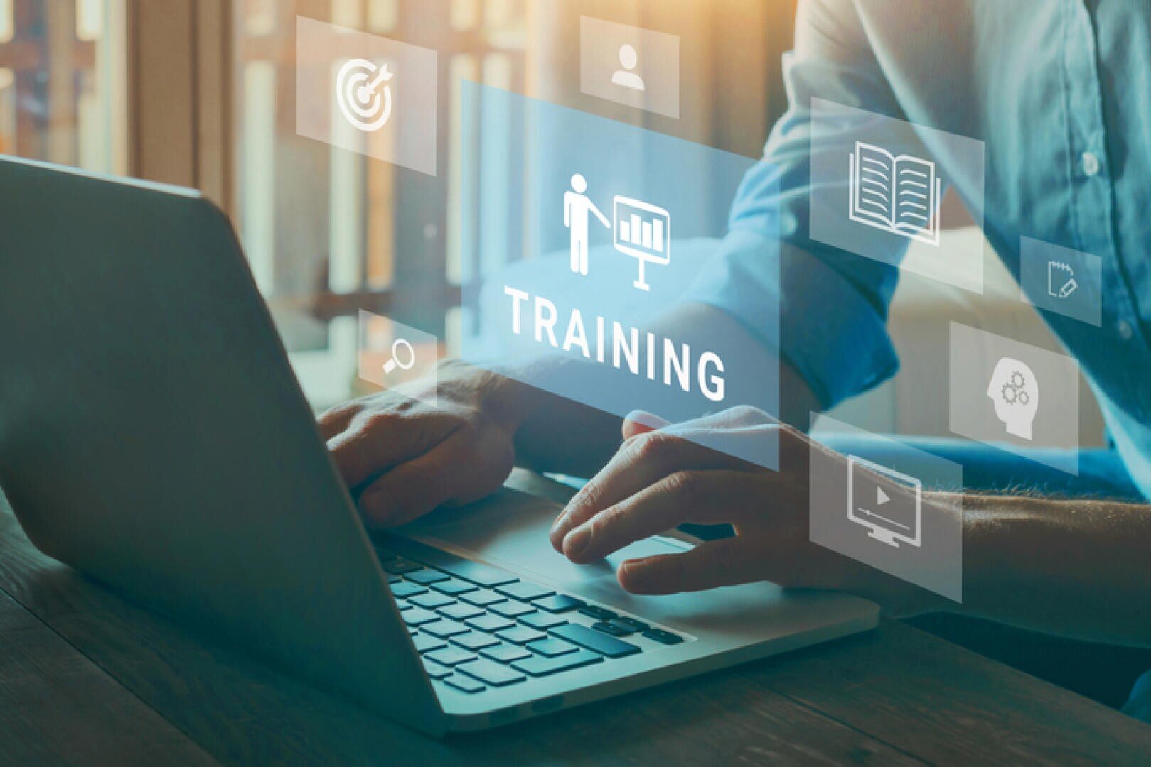 Eine Person tippt auf einem Laptop mit virtuellen Symbolen, die über der Tastatur schweben, darunter das Wort TRAINING und Symbole für Ziele, Bücher und Online-Lernen, was auf Online-Ausbildung oder berufliche Weiterbildung hindeutet.