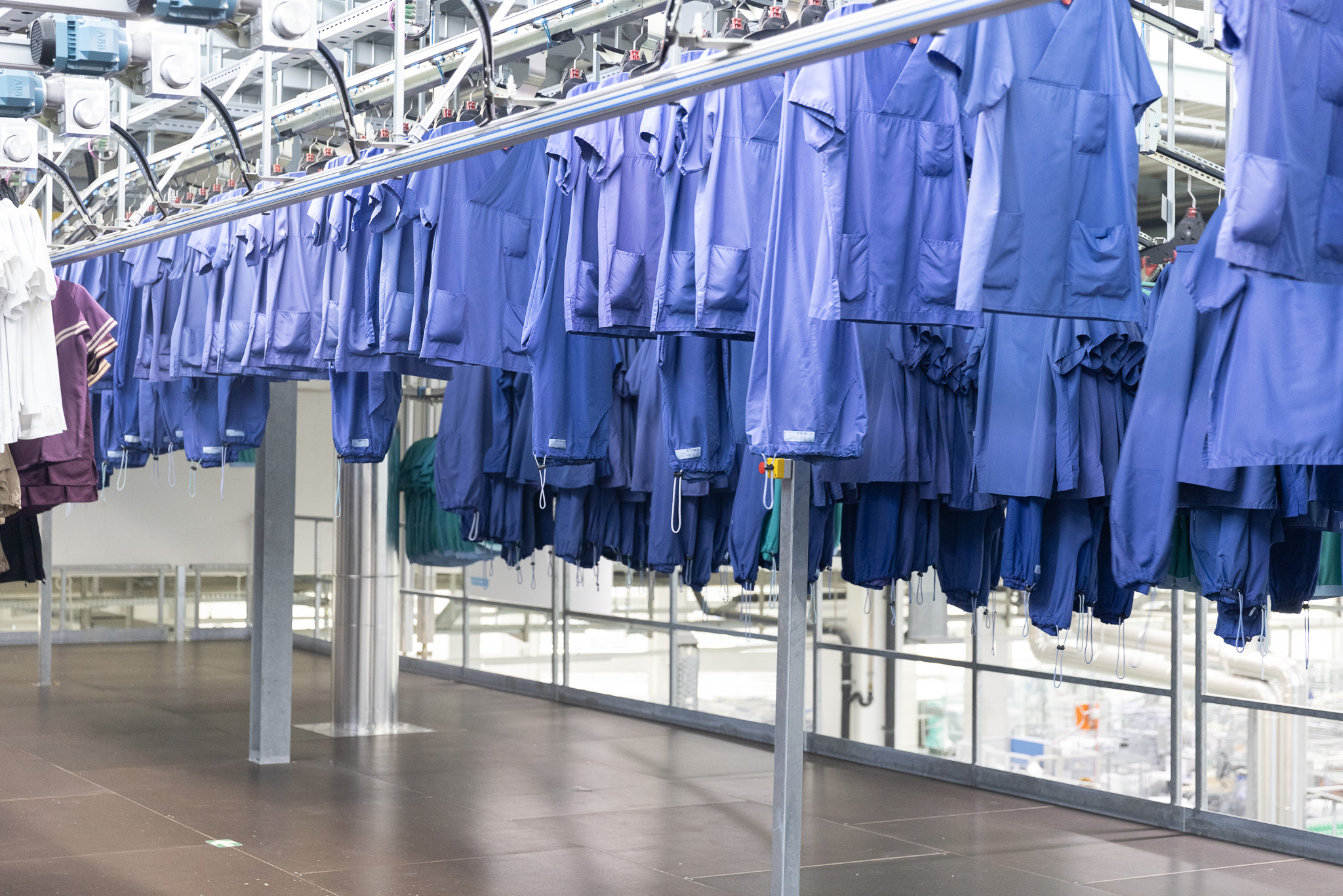 Des tenues chirurgicales bleues suspendues à un système de convoyage automatisé dans une blanchisserie, avec en arrière-plan un éclairage vif et de grandes fenêtres.
