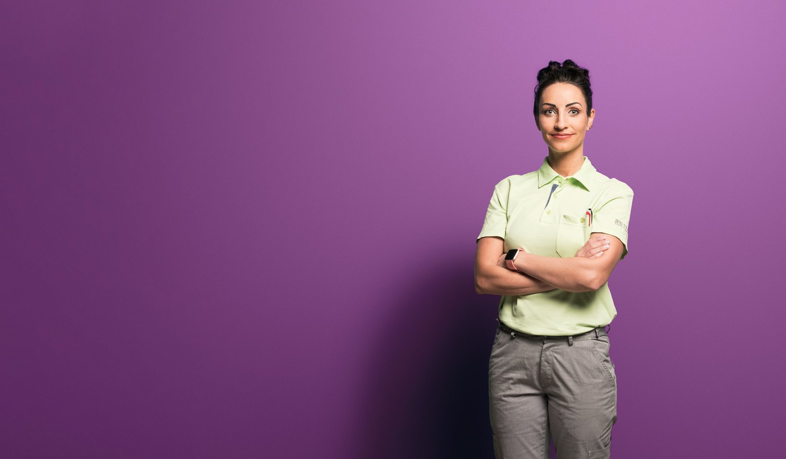 Une femme portant un polo vert clair et un pantalon gris se tient debout, les bras croisés, souriante, devant un fond violet uni.