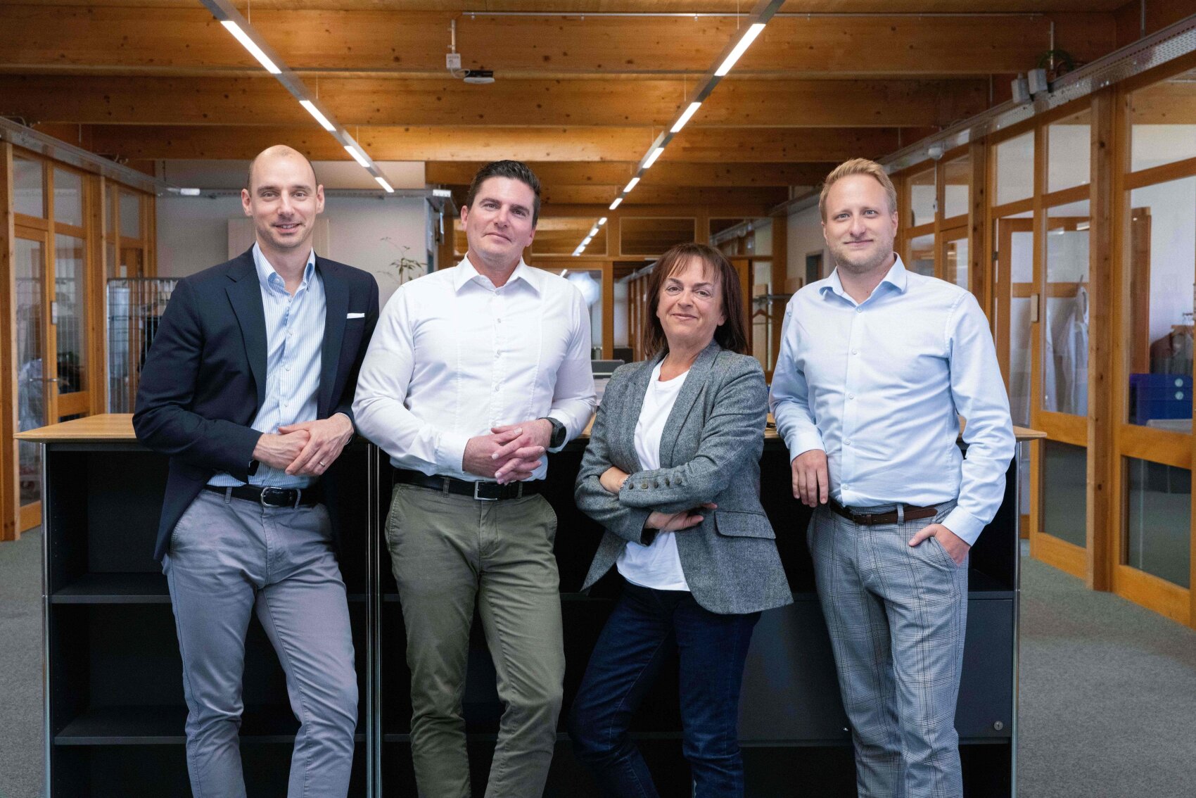 Quatre personnes – trois hommes et une femme – se tiennent ensemble dans un bureau aux poutres apparentes et aux cloisons vitrées ; elles prennent la pose et sourient à la caméra. Vêtues d'une tenue « business casual », elles sont adossées à des étagères noires.